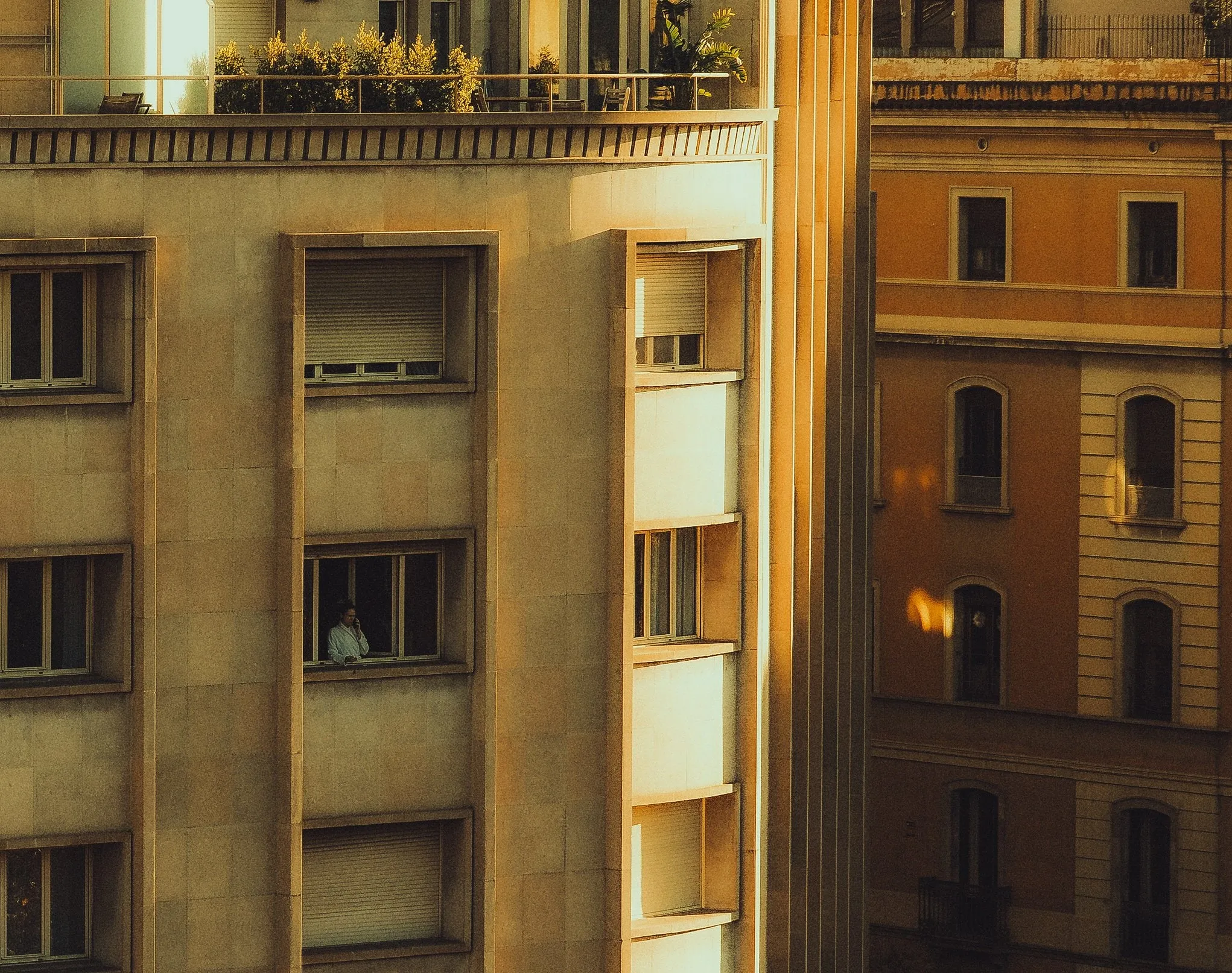 An apartment building in low golden sunlight, with a person standing at one window