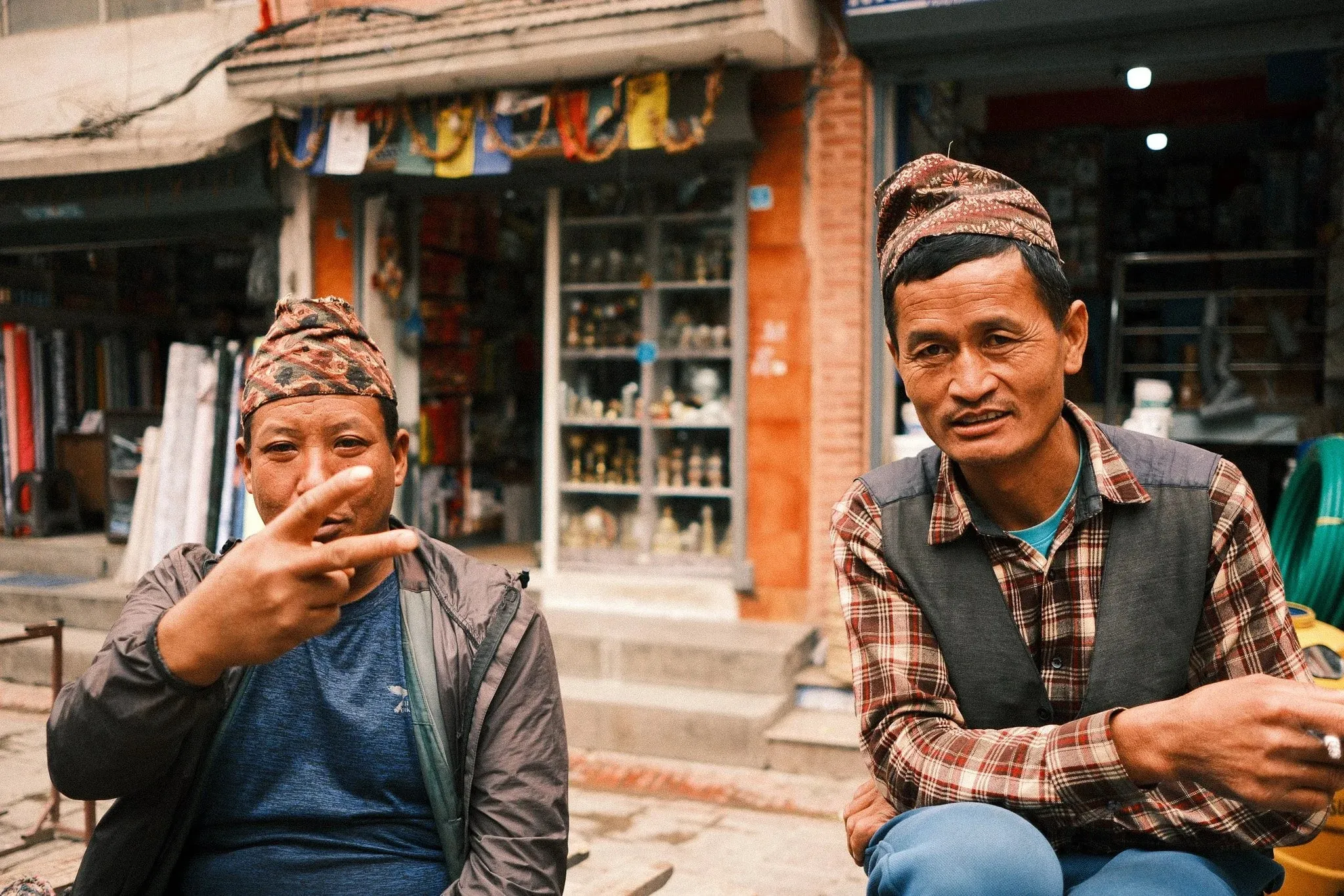 Two men in dhaka topi hats sitting outside a shop, one making a peace sign