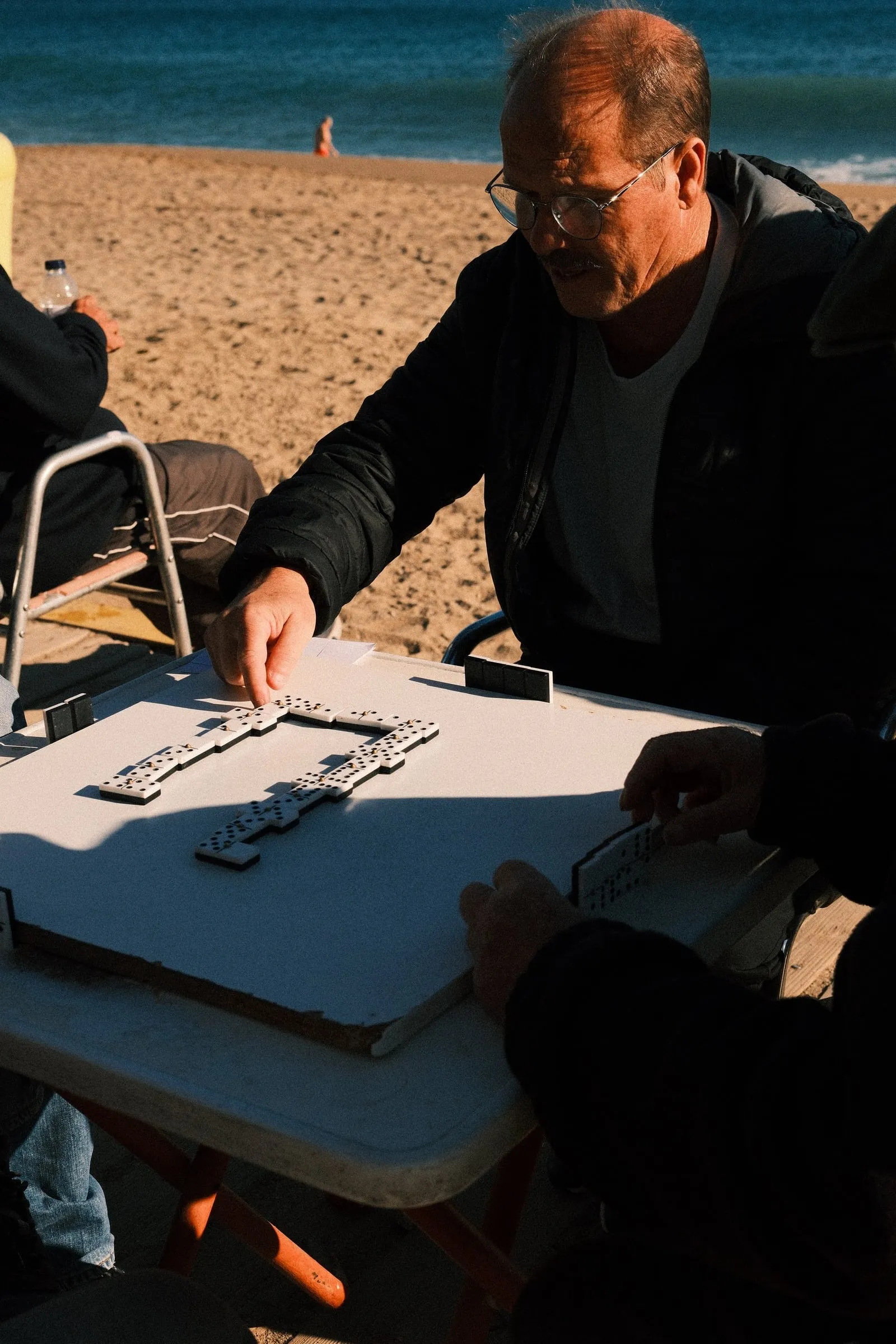 A man lays a domino on a table on the sand at Barceloneta Beach