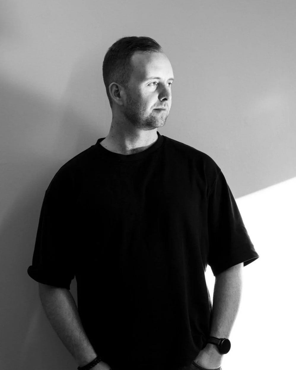Black-and-white portrait of Ben Arvi in a black T-shirt, leaning against a wall and looking to the side