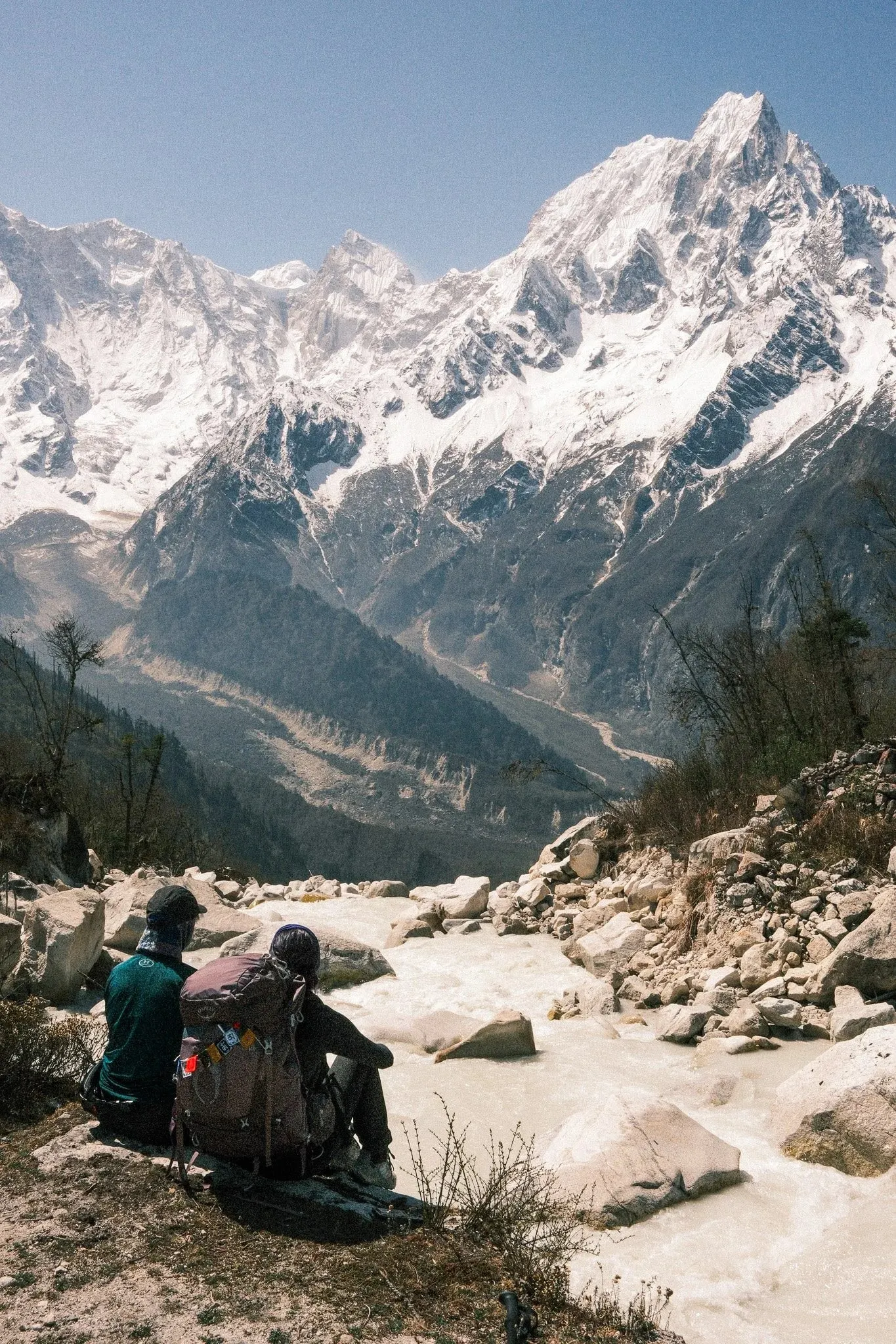 Two trekkers resting on rocks beside a glacial river below snow peaks near Bimthang