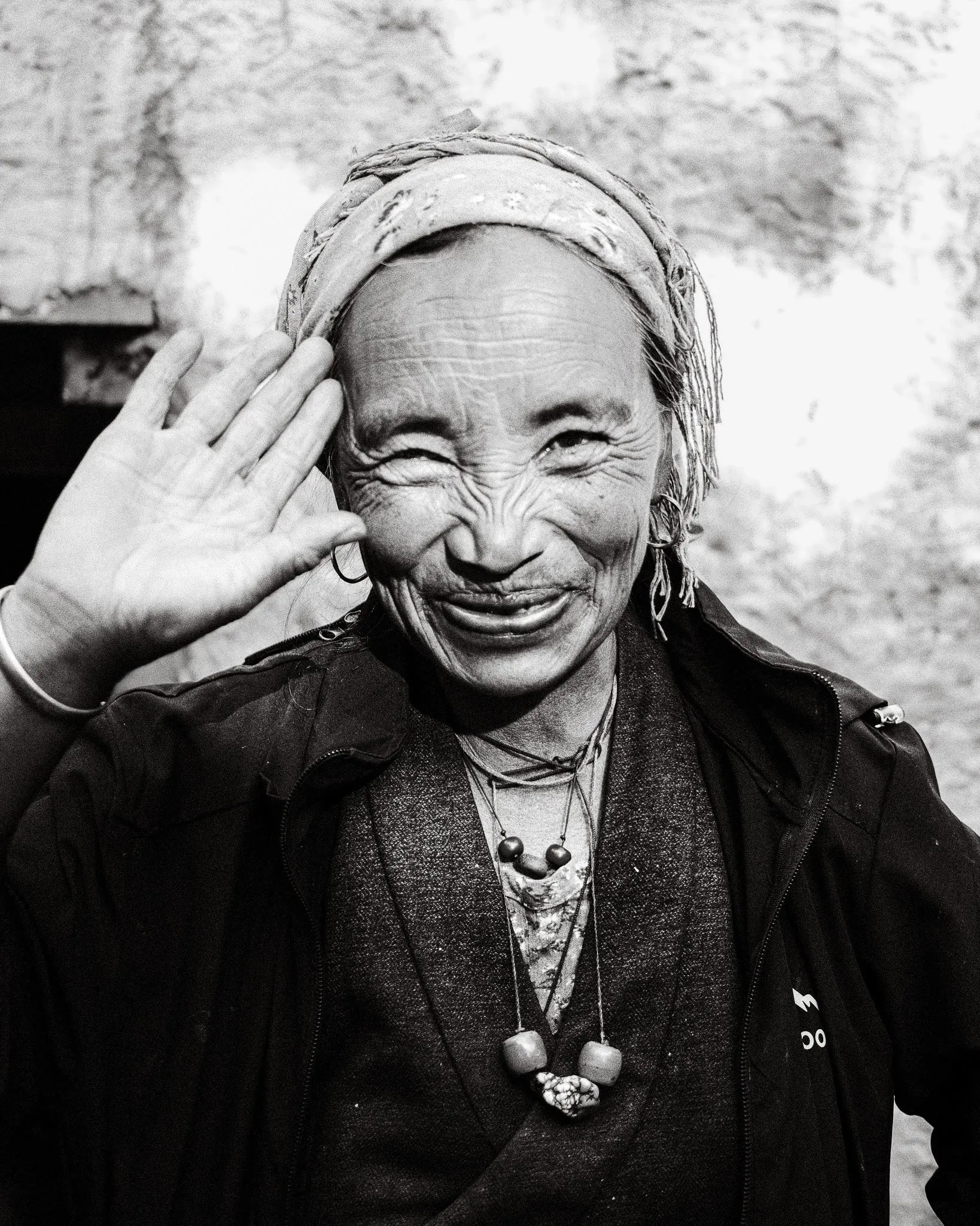 Black and white portrait of a smiling older woman wearing a headscarf and raising her hand in Dhakmar, Upper Mustang, Nepal.