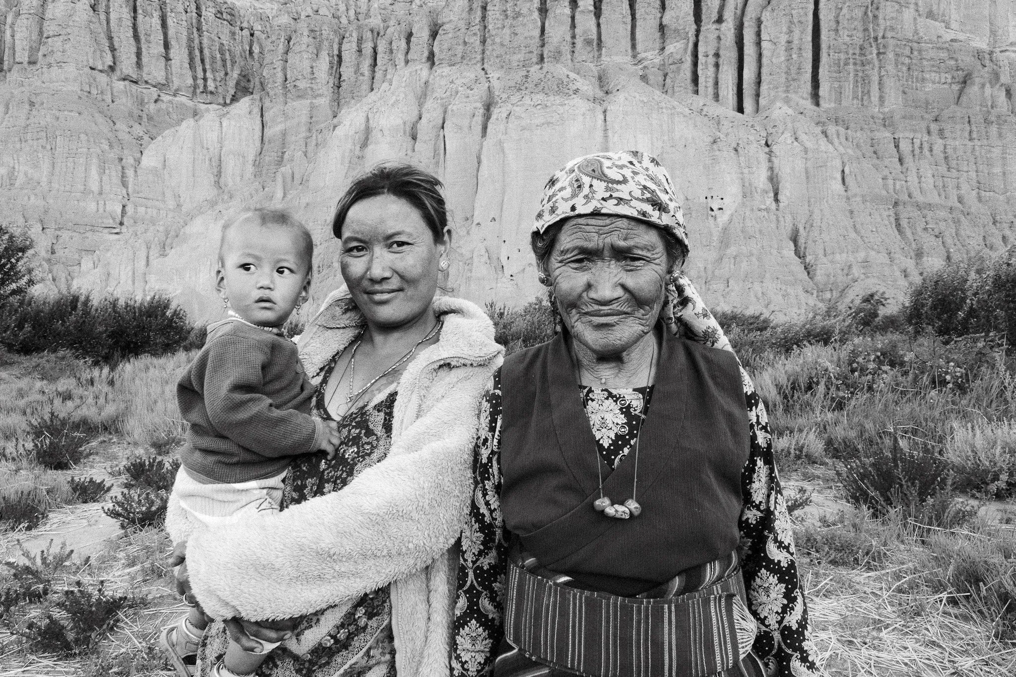 Black and white portrait of three generations of a family, with a woman holding a child beside an older woman beneath the cliffs in Chuksang, Upper Mustang, Nepal.