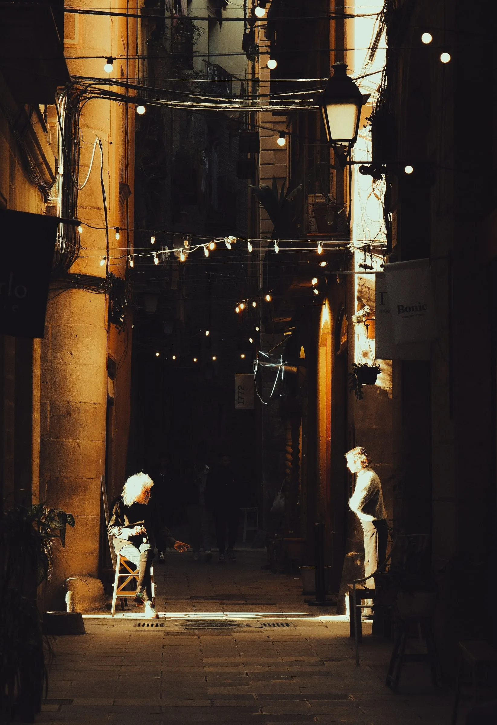 An older woman sits on a chair in a shaft of sunlight in a narrow alley hung with string lights