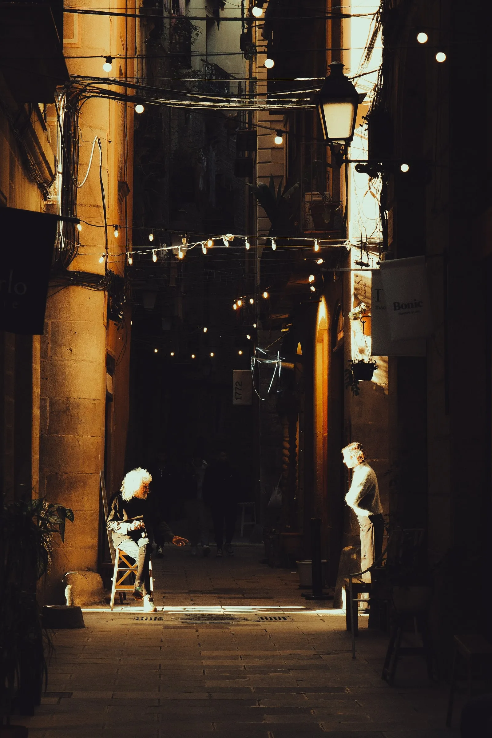 An older woman sits on a chair in a shaft of sunlight in a narrow alley hung with string lights
