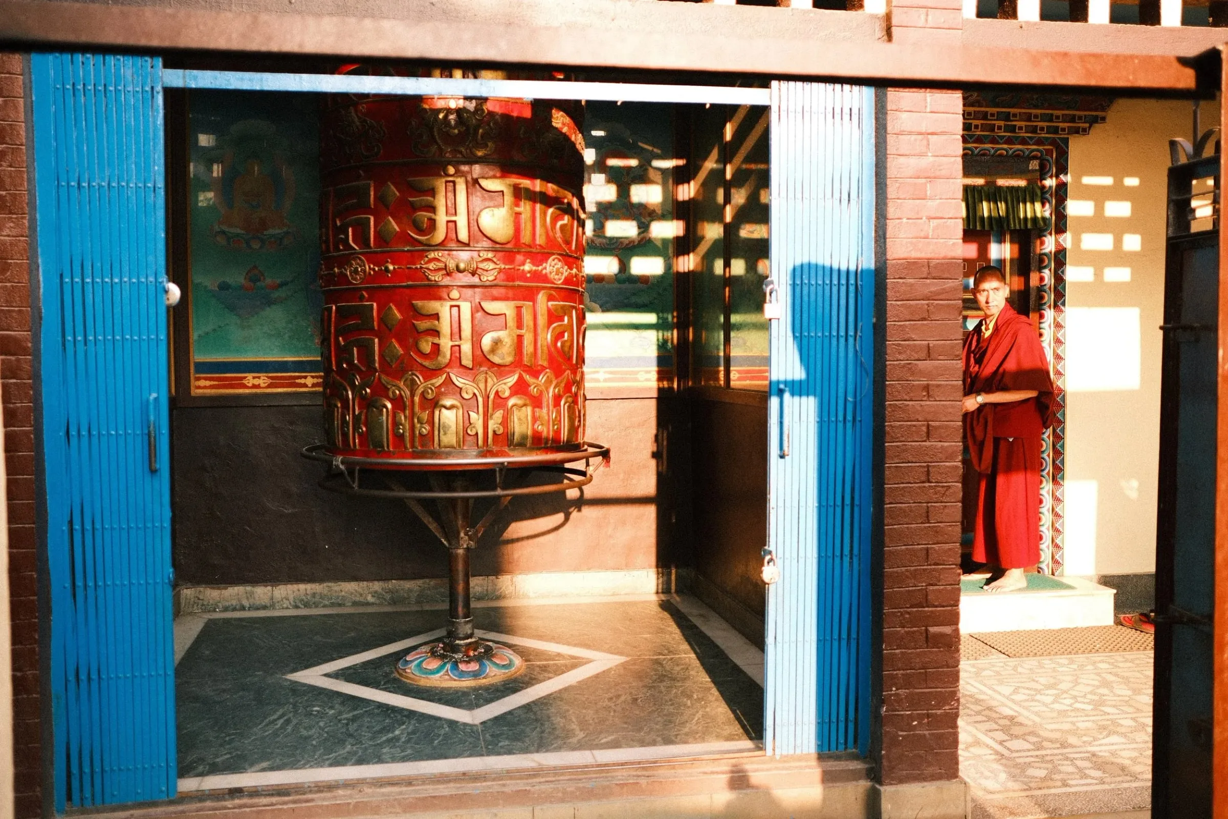 A large red prayer wheel seen through a blue doorway at Boudhanath, with a monk to one side