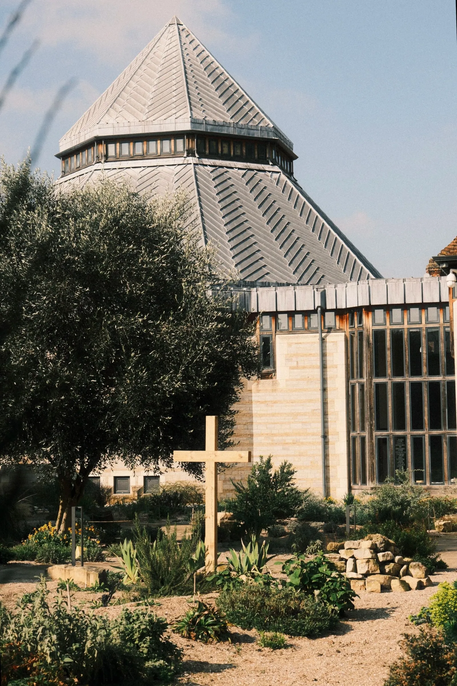 A wooden cross in a garden of shrubs in front of a modern building with a grey pointed roof