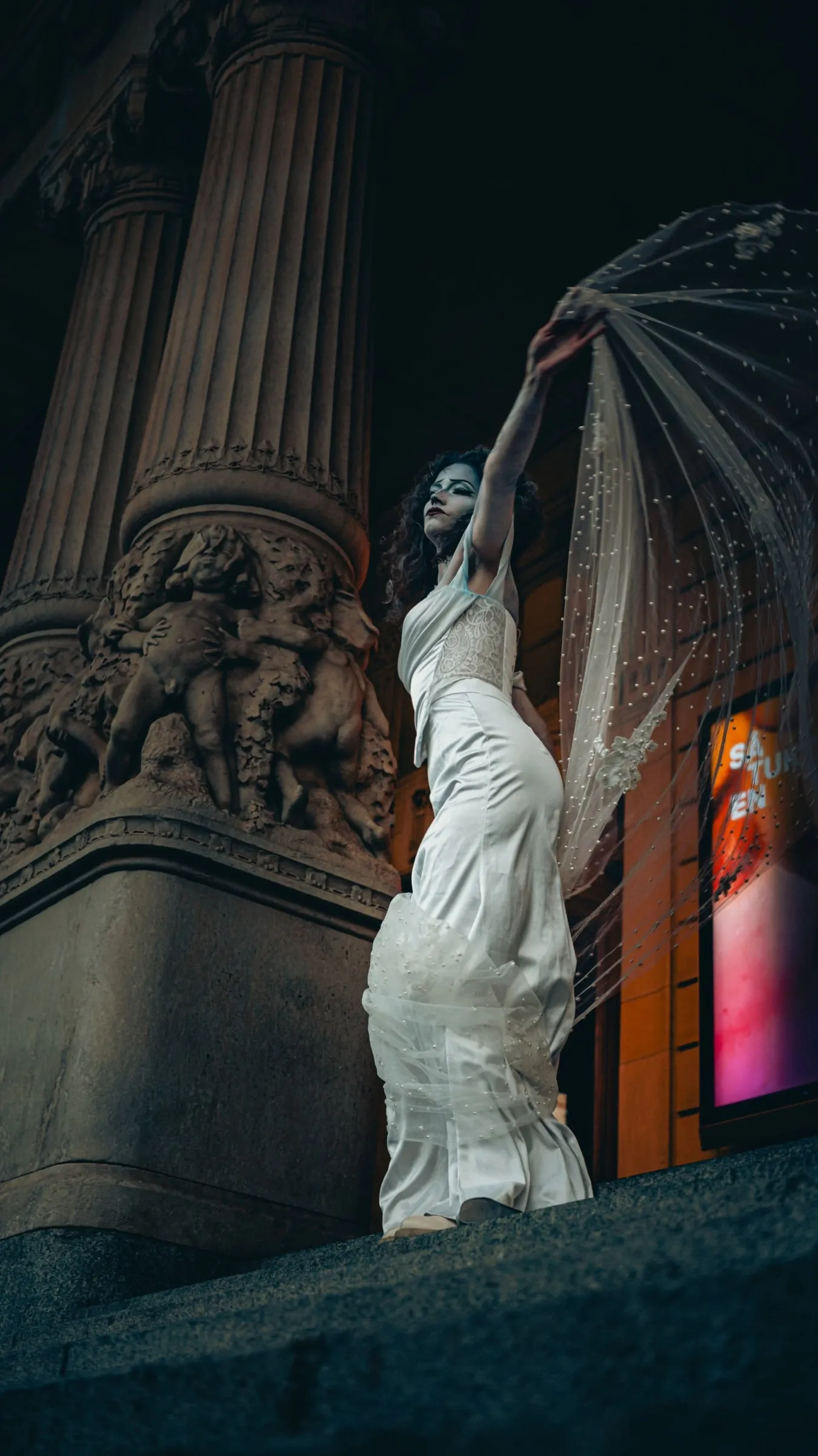 A woman in a white dress and veil posing beside carved stone columns at the theatre
