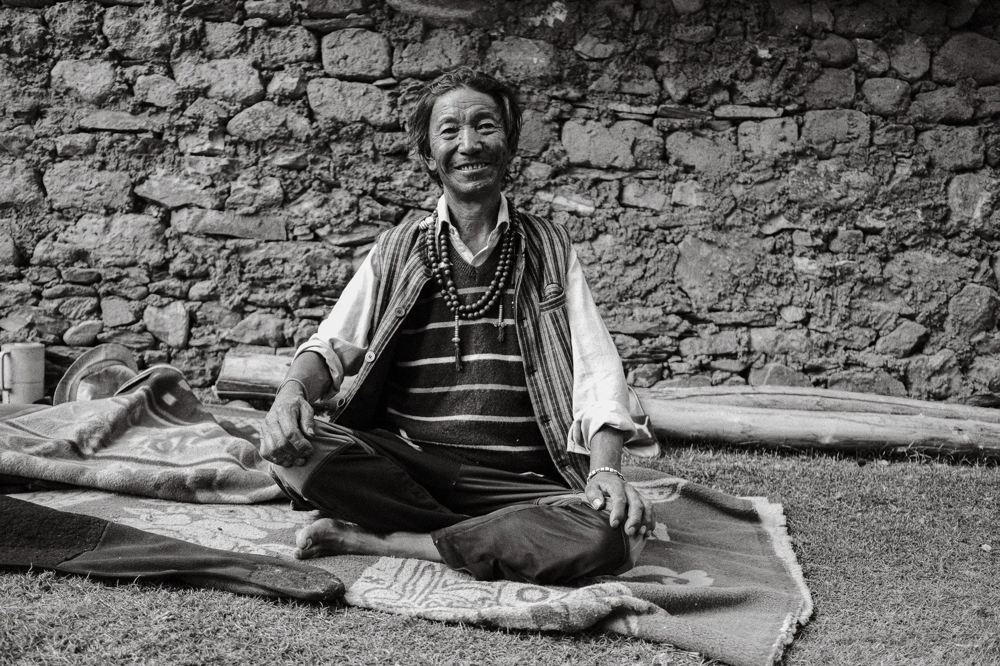 Black and white photo of a smiling man sitting cross-legged on a blanket in front of a stone wall in Dhakmar
