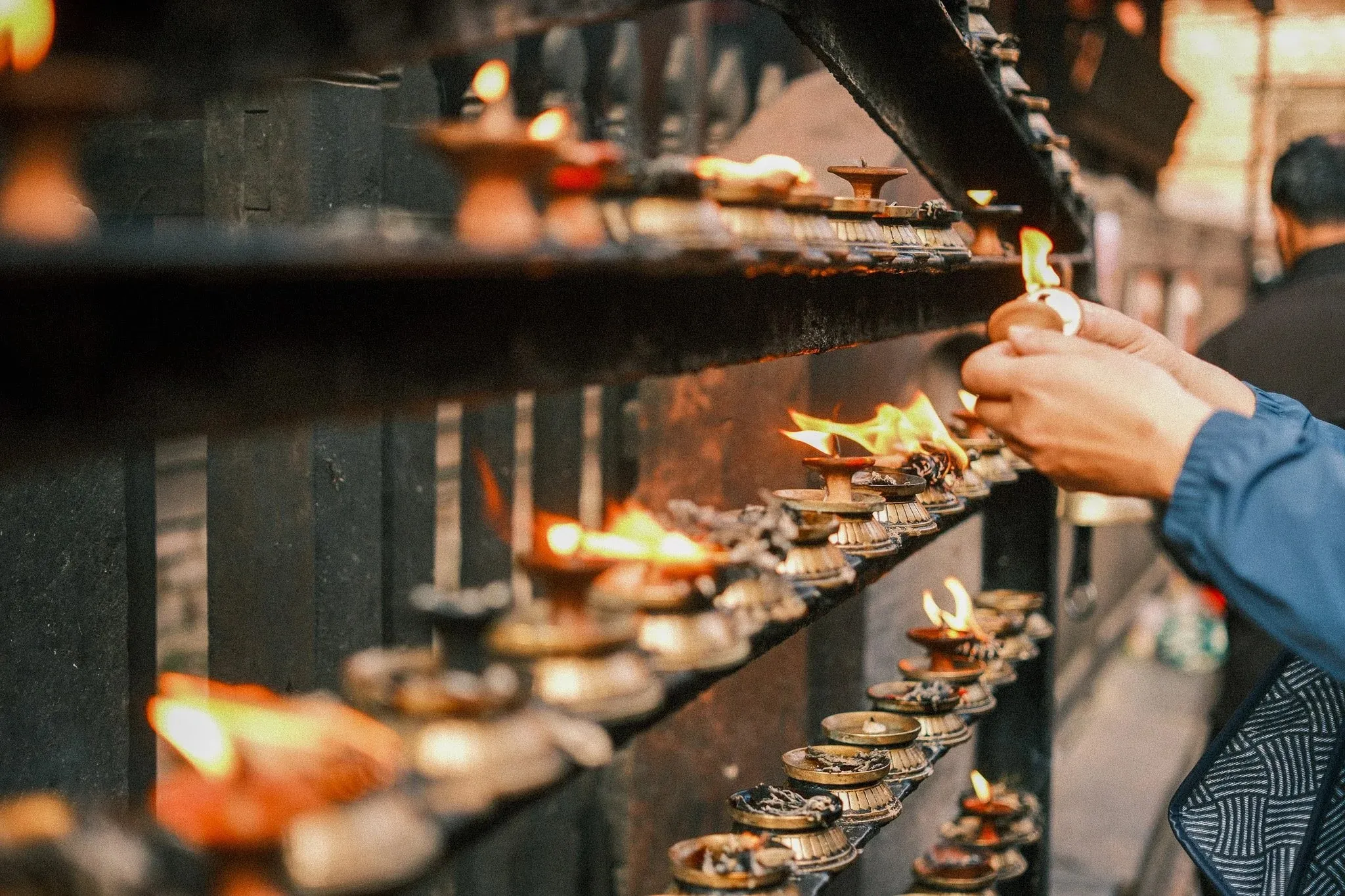 A hand lighting a row of butter lamps on a shrine rack in Kathmandu Durbar Square