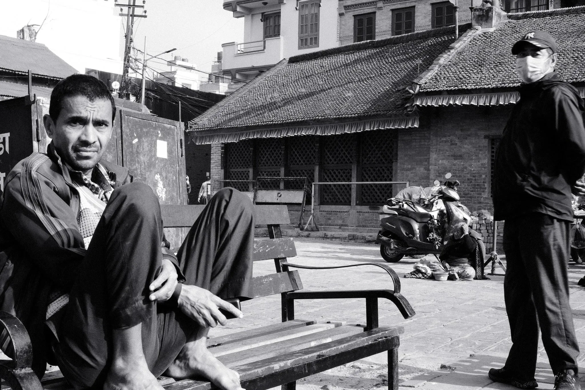 Black and white photo of a man sitting on a bench, with another man standing nearby
