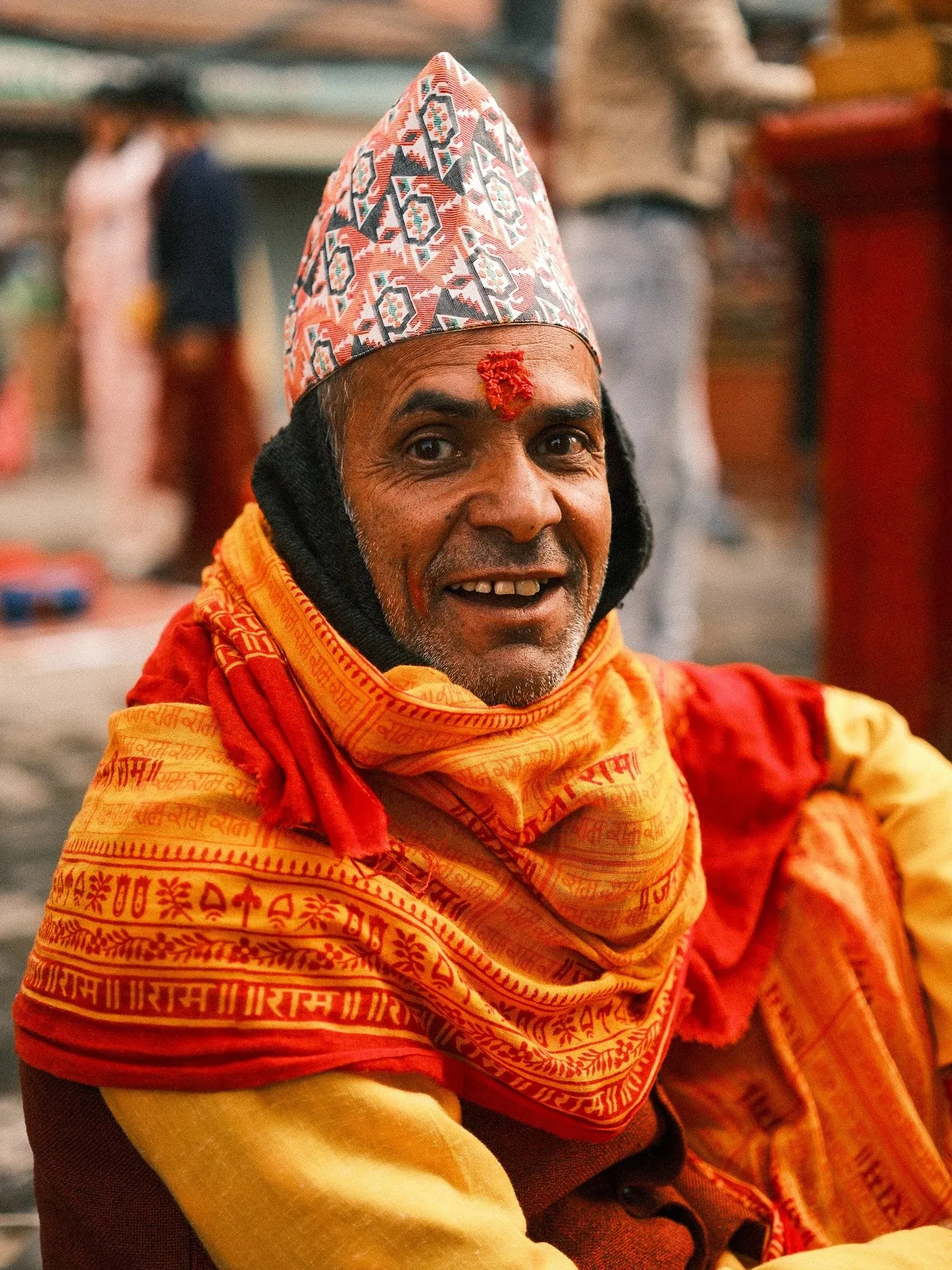A smiling man in a dhaka topi and an orange scarf printed with lettering
