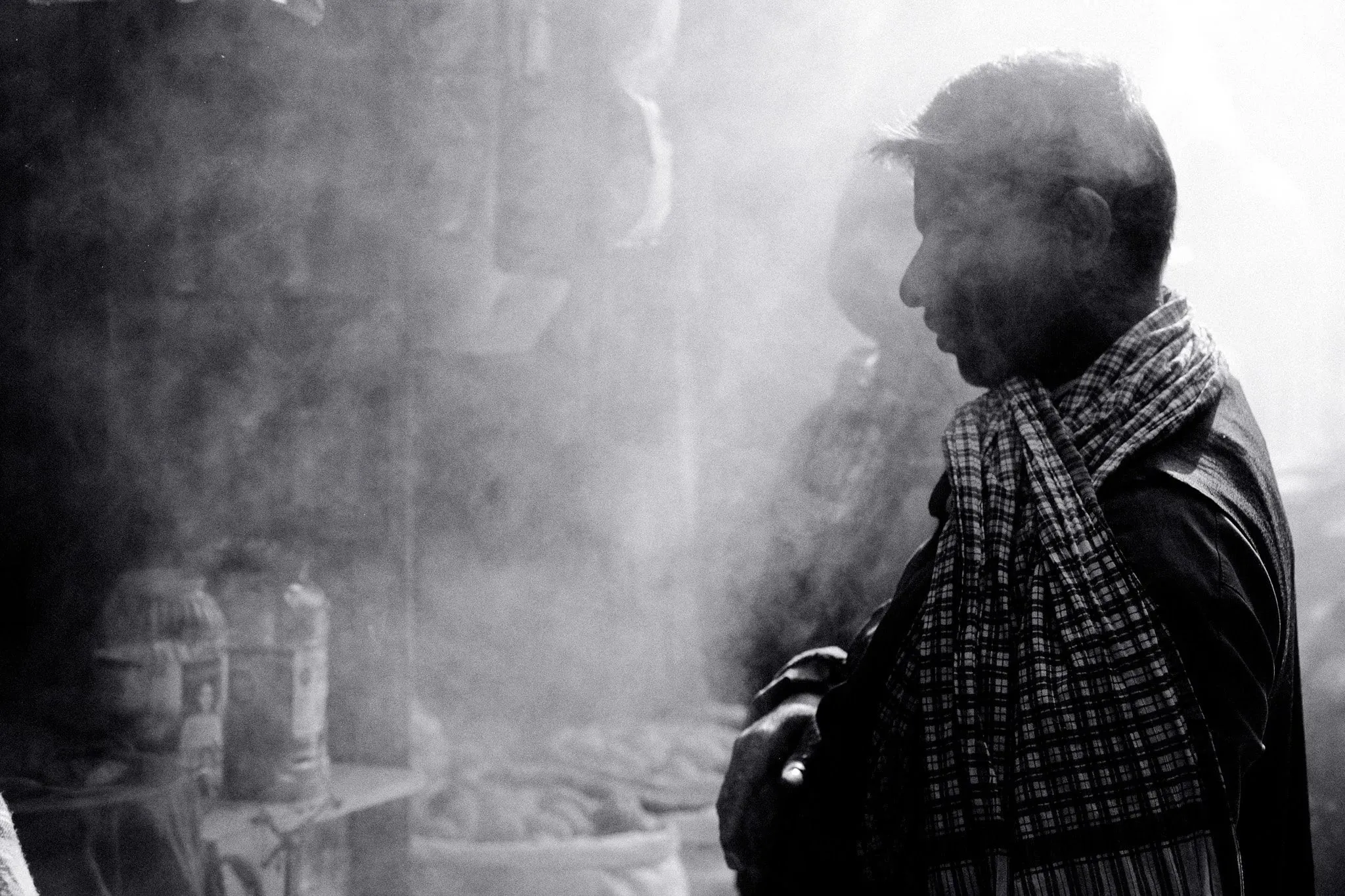 Black and white photo of a man in a checked scarf standing in incense smoke