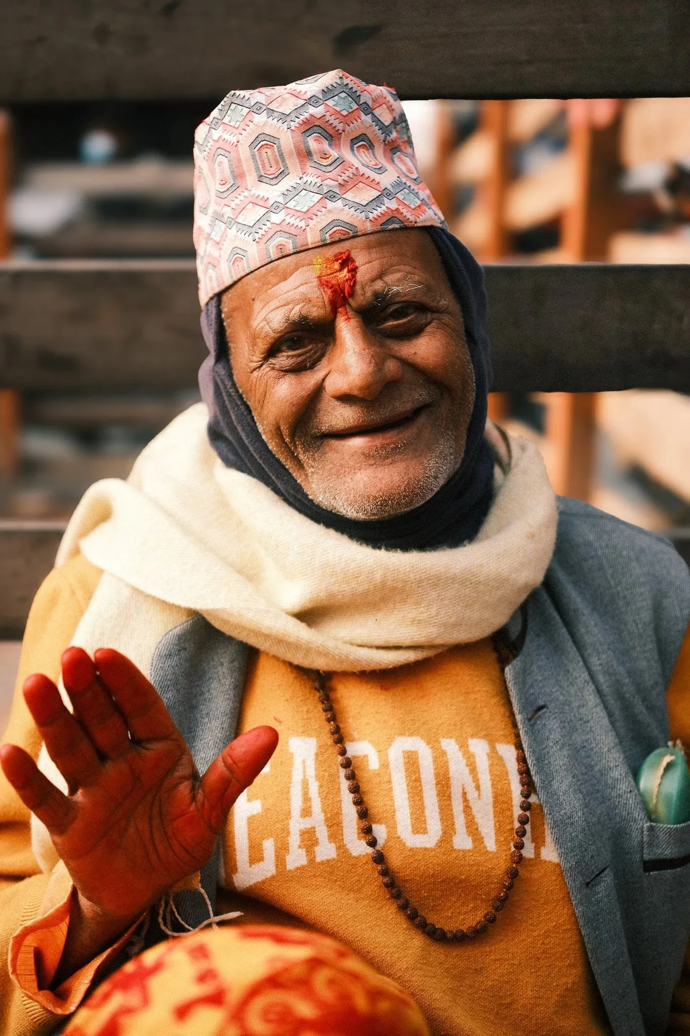 A smiling older man in a dhaka topi with tika on his forehead raises his hand