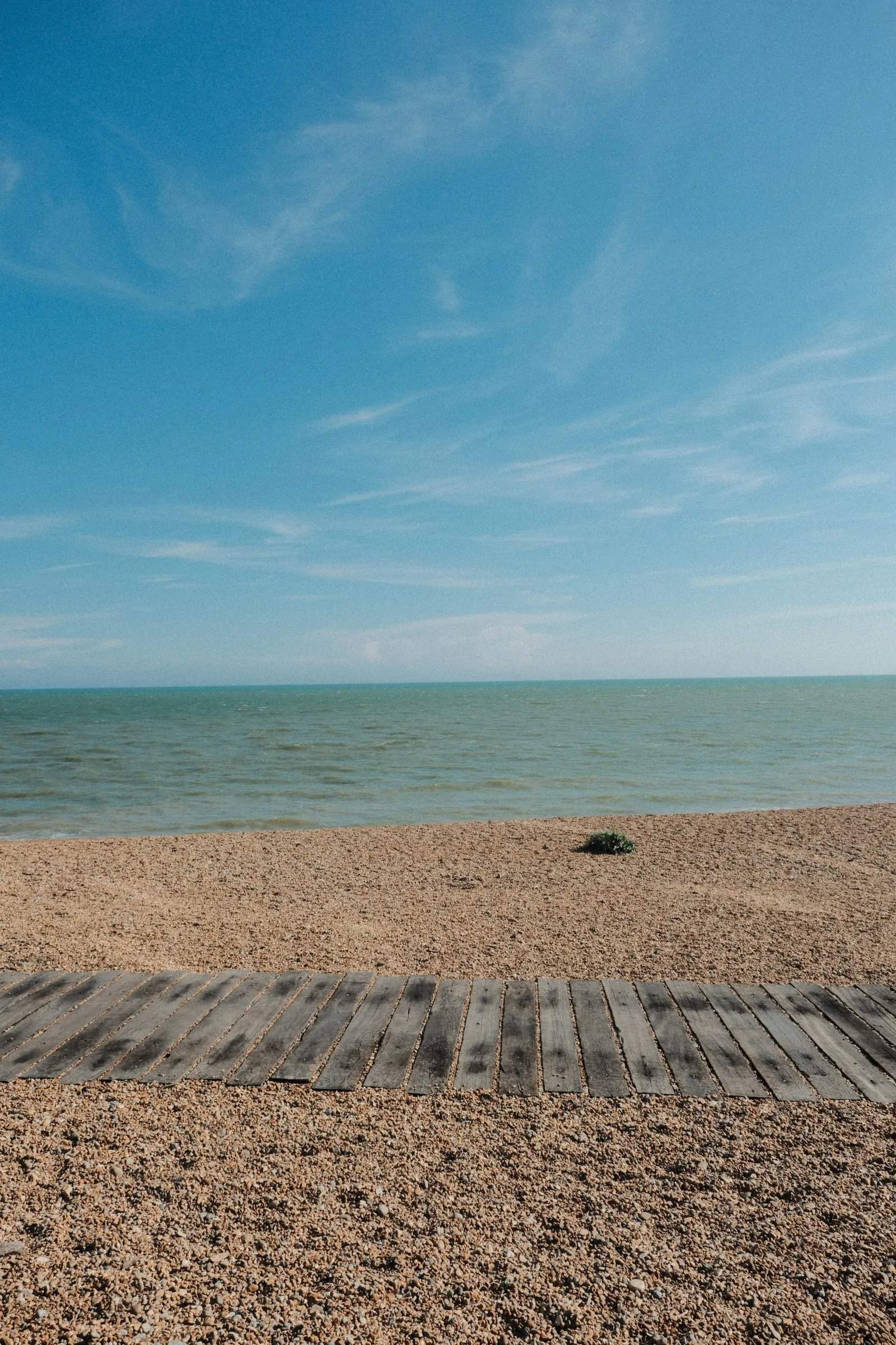 A wooden boardwalk across the shingle towards a calm sea under a blue sky