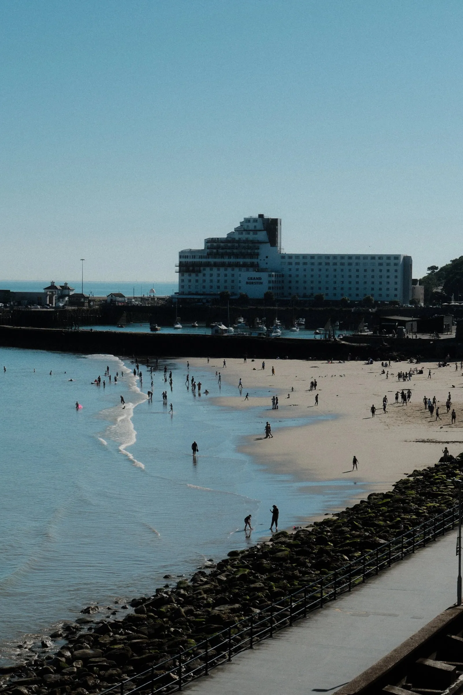 People on the beach at the water’s edge, with the harbour and a large hotel behind