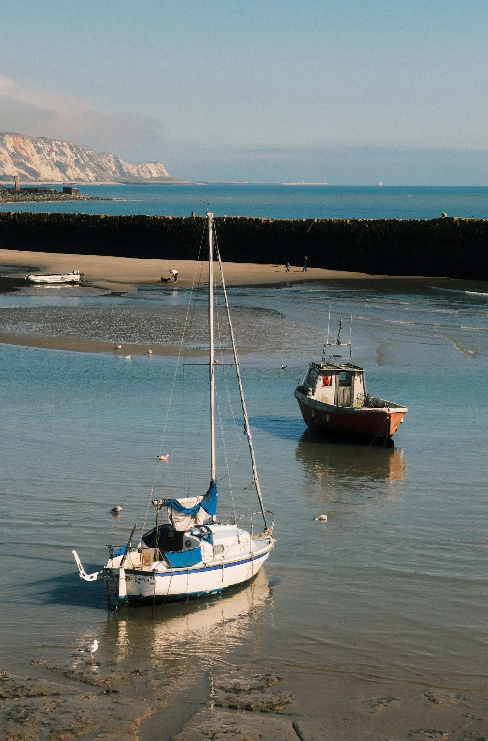 A sailing boat and a fishing boat at low tide in the harbour, with white cliffs in the distance