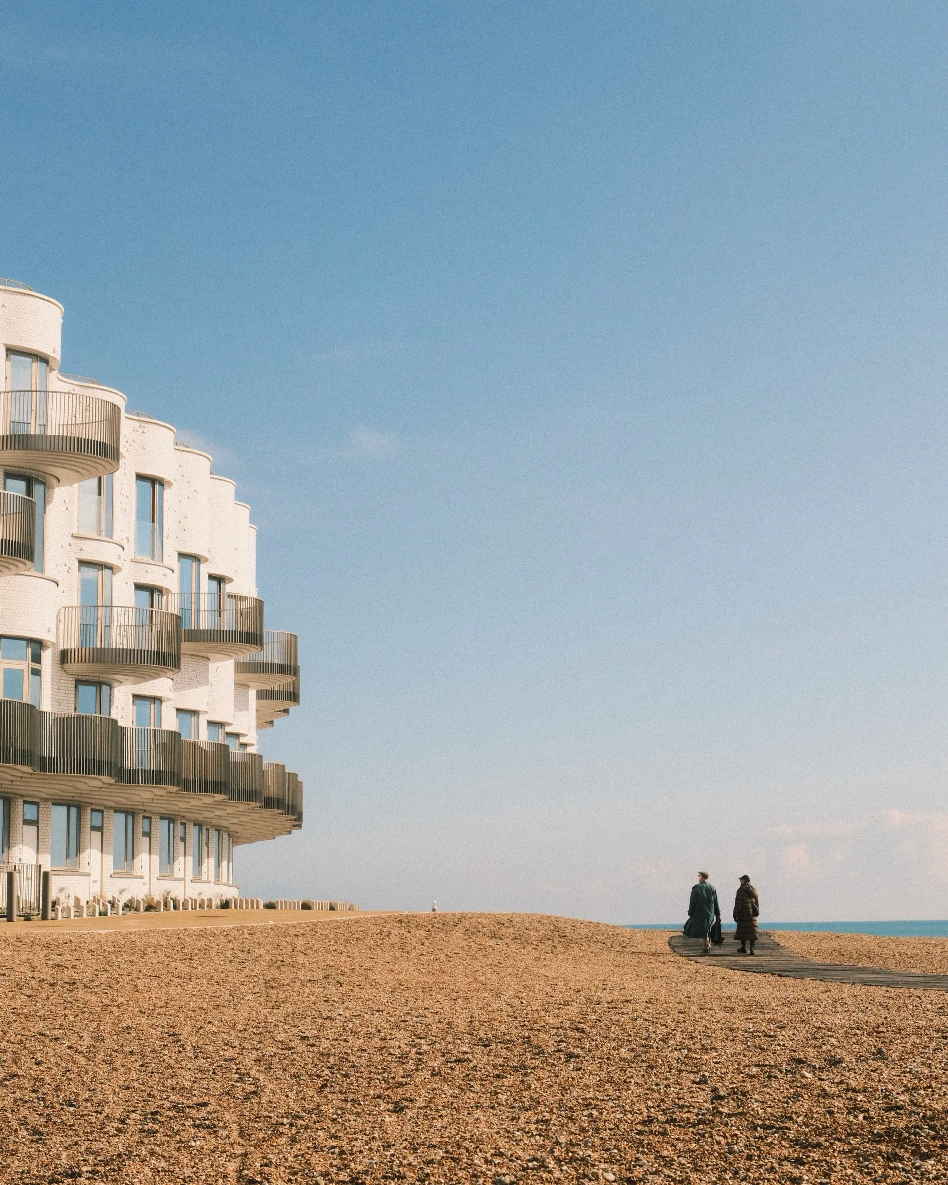 Two people walking on the shingle beside curved white seafront apartments