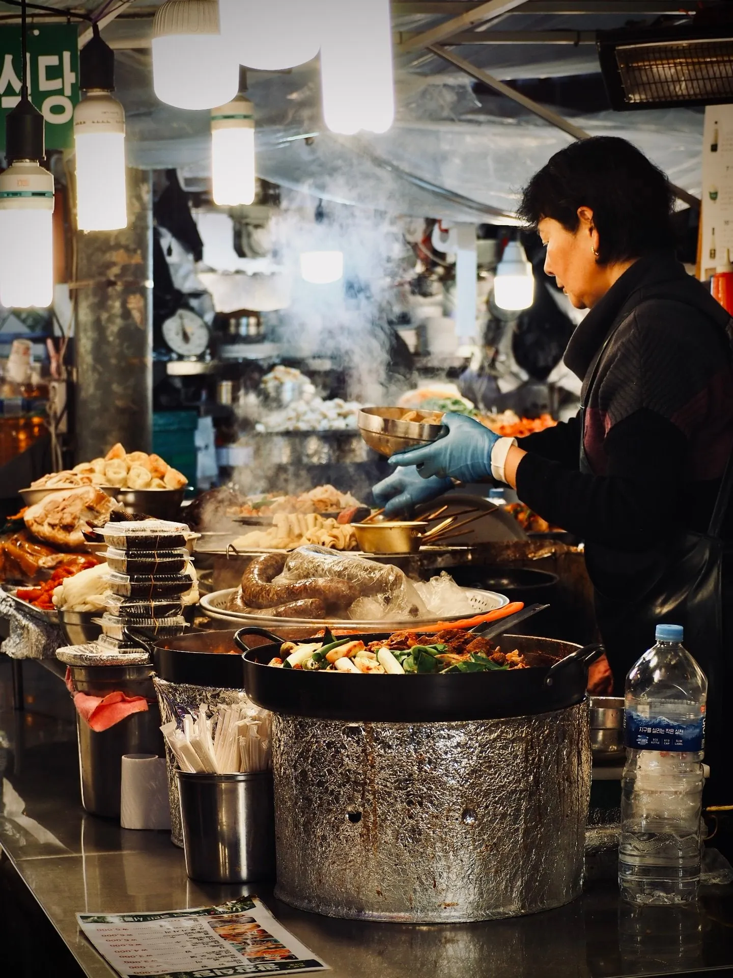 A cook in blue gloves works behind steaming pots and dishes at Gwangjang Market