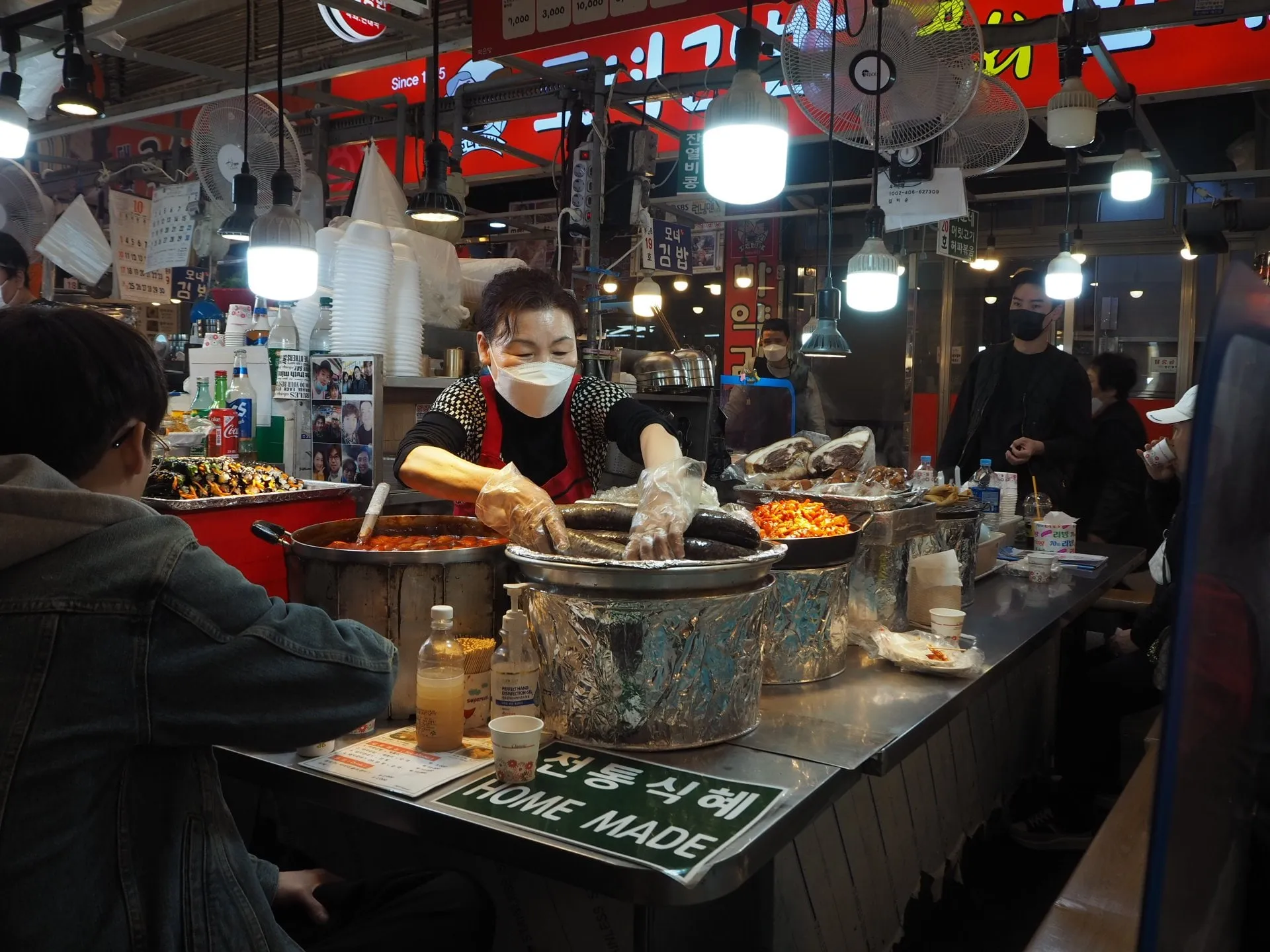 A vendor in a mask and gloves prepares food at a busy stall in Gwangjang Market