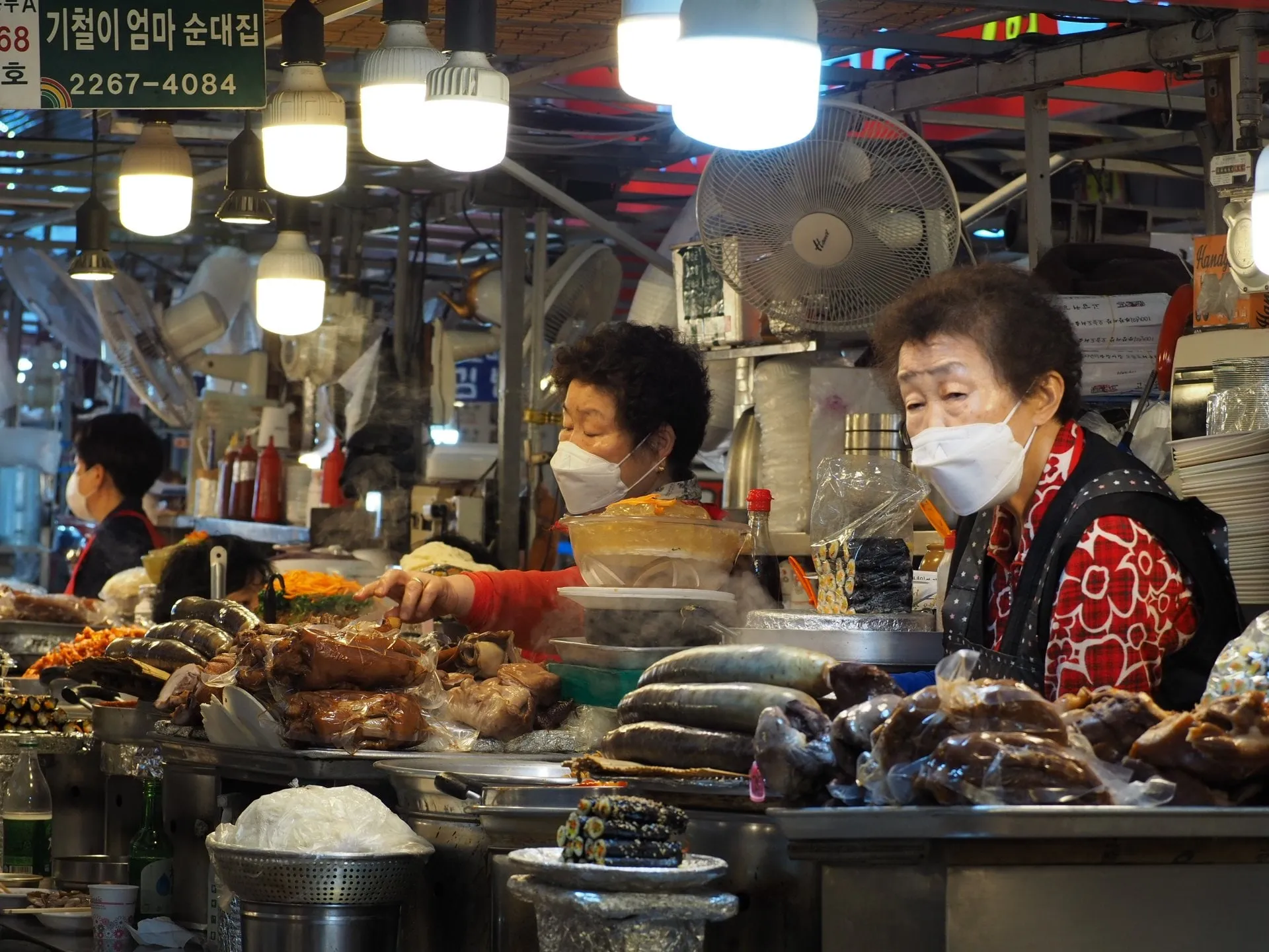 Vendors in masks behind a counter piled with food under hanging lights at Gwangjang Market
