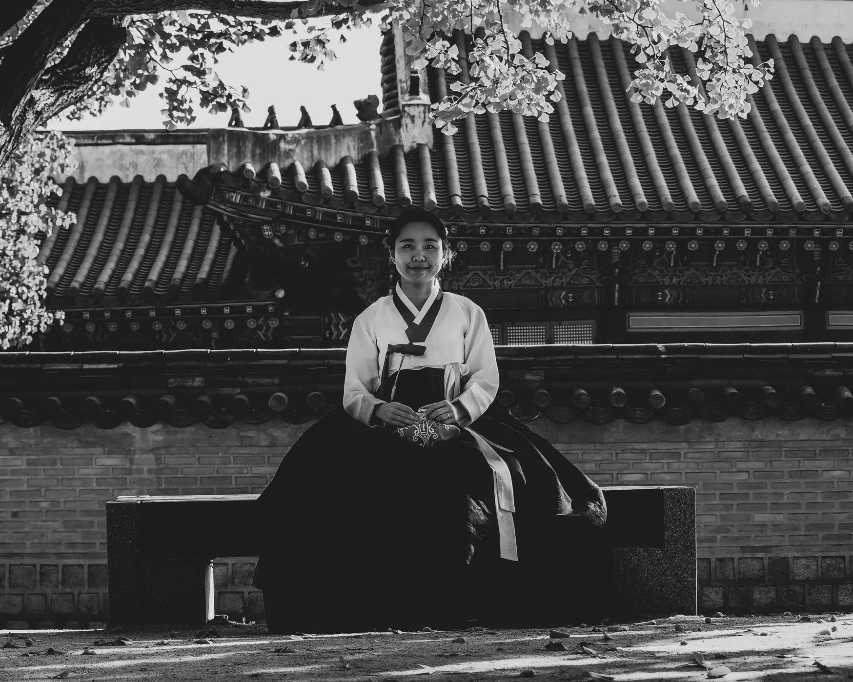 Black and white photo of a young woman in hanbok sitting on a bench in front of palace buildings
