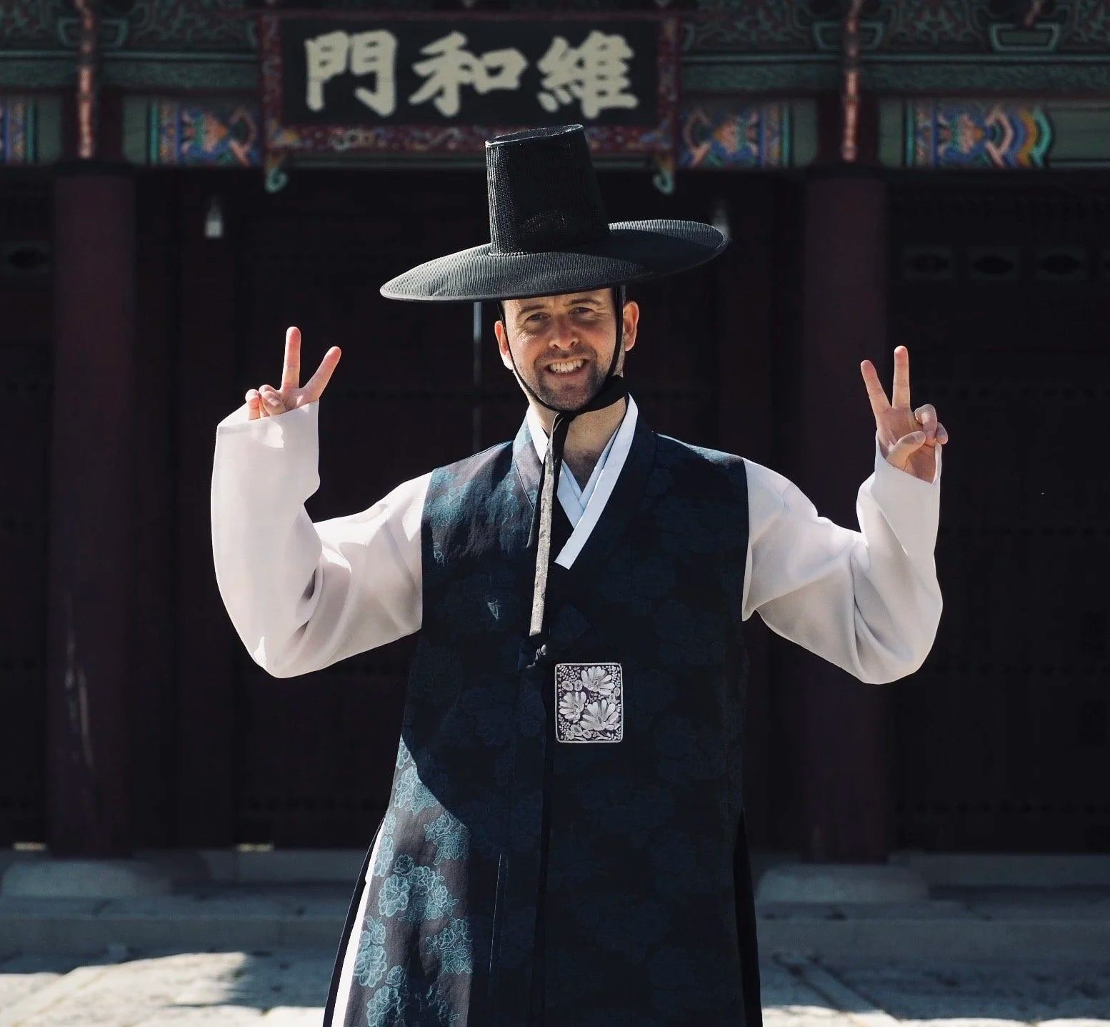 A man in a hanbok and a black gat hat makes peace signs in front of a palace gate in Seoul