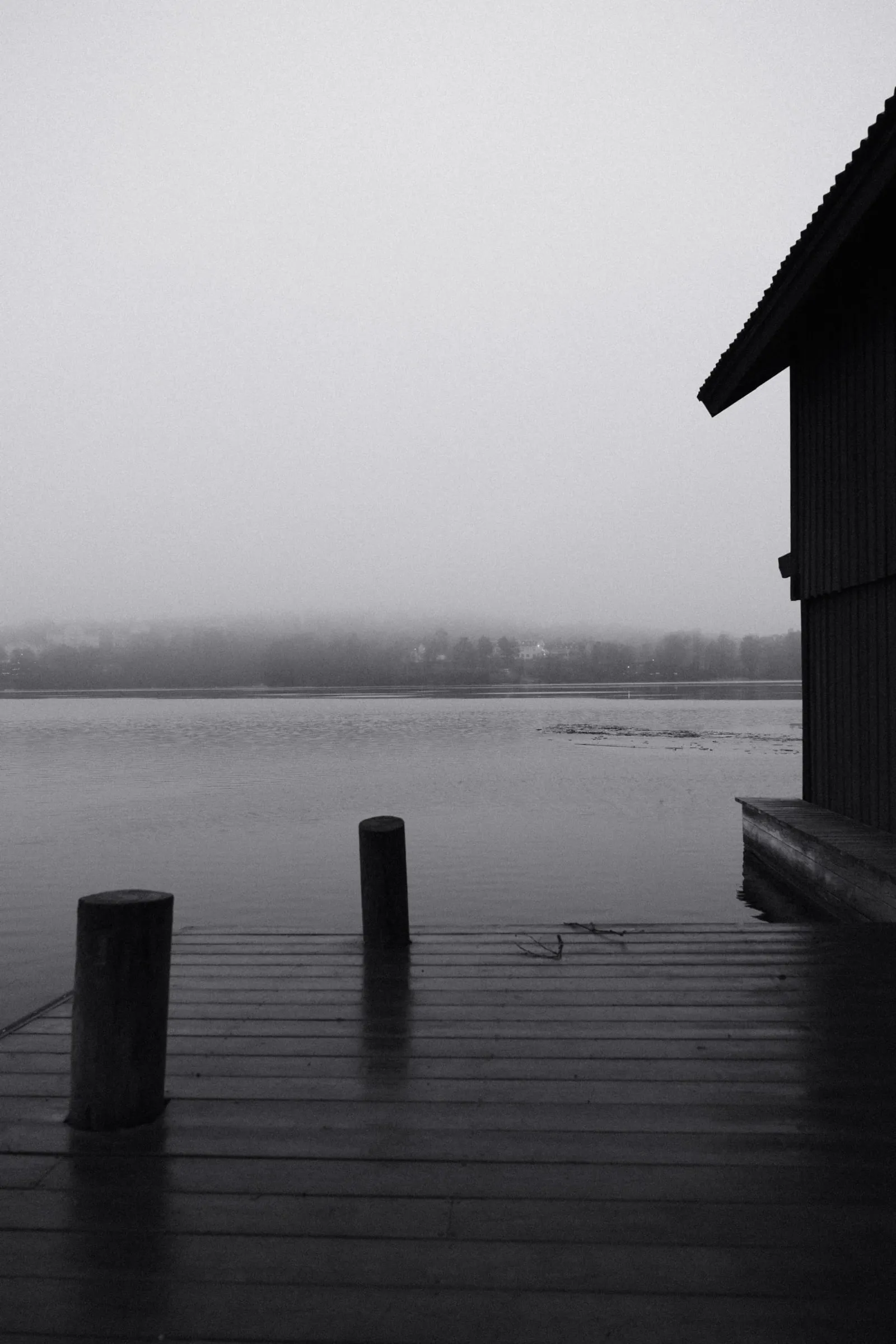 Black and white photo of wooden posts on a jetty beside a boathouse, looking out over misty water