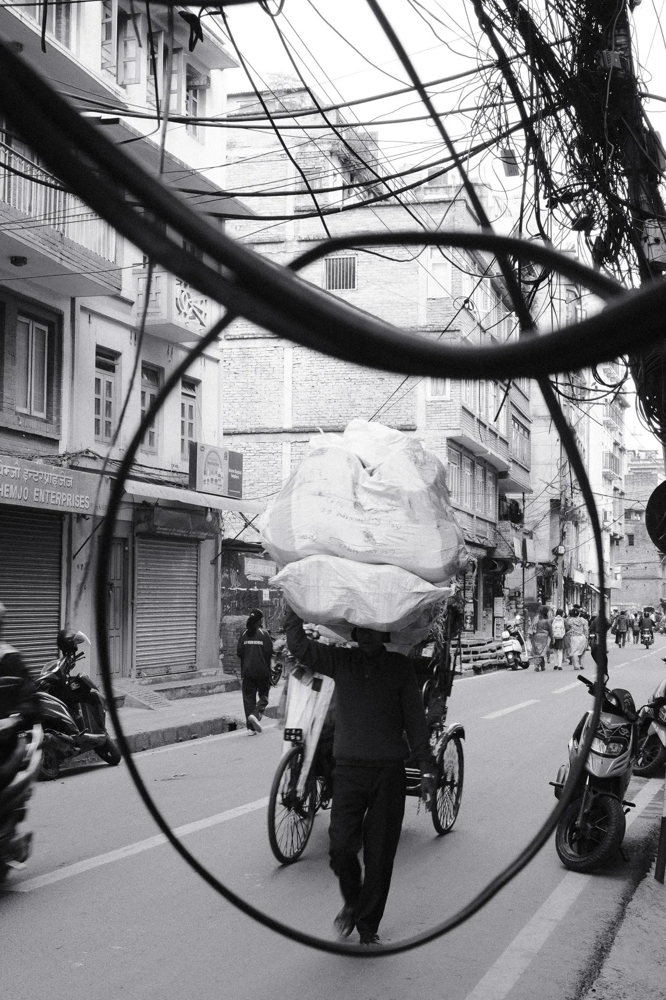 Black and white photo of a cycle rickshaw with a large load framed by a loop of overhead wire