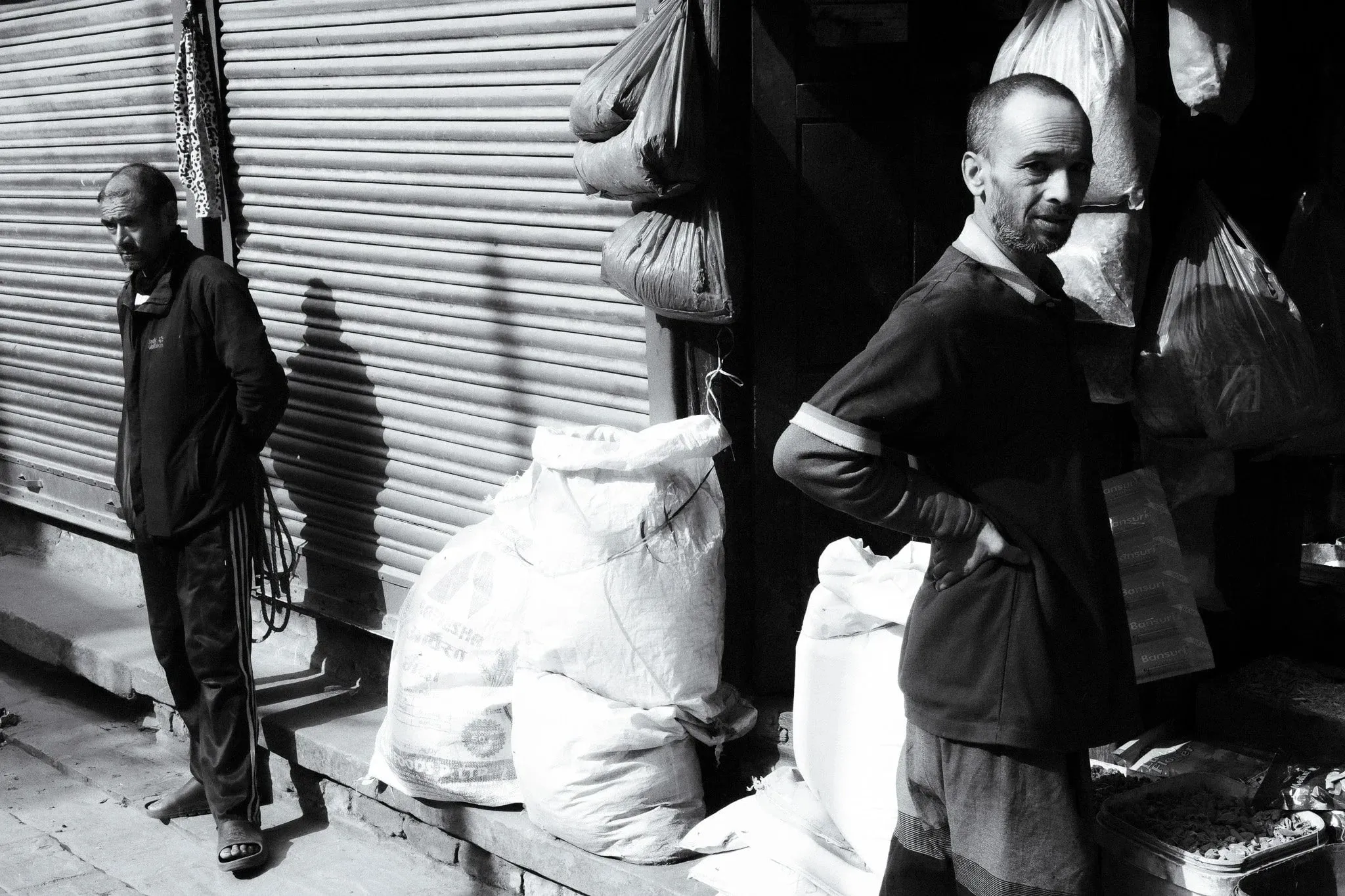Black and white photo of two men standing by sacks outside a shop with a shuttered front