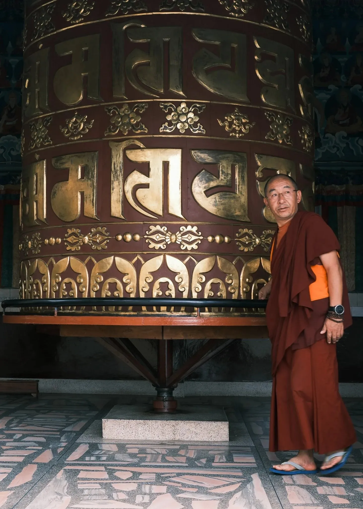 A monk standing beside a large golden prayer wheel at Kopan Monastery