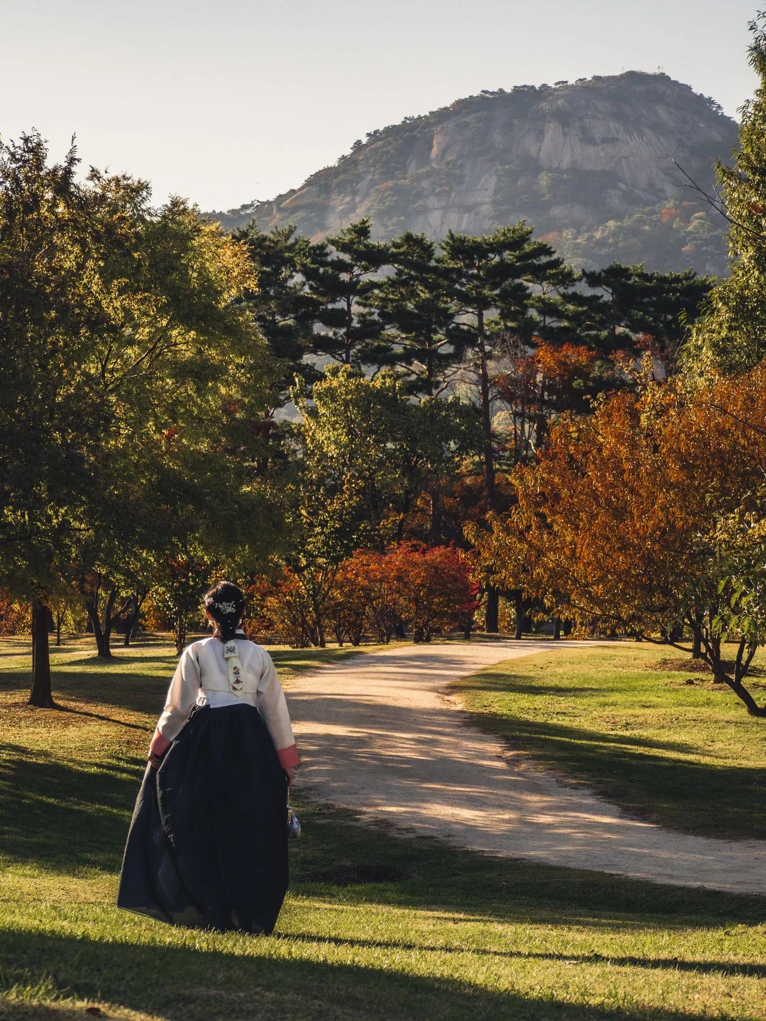 Woman in hanbok walking along a path among autumn trees