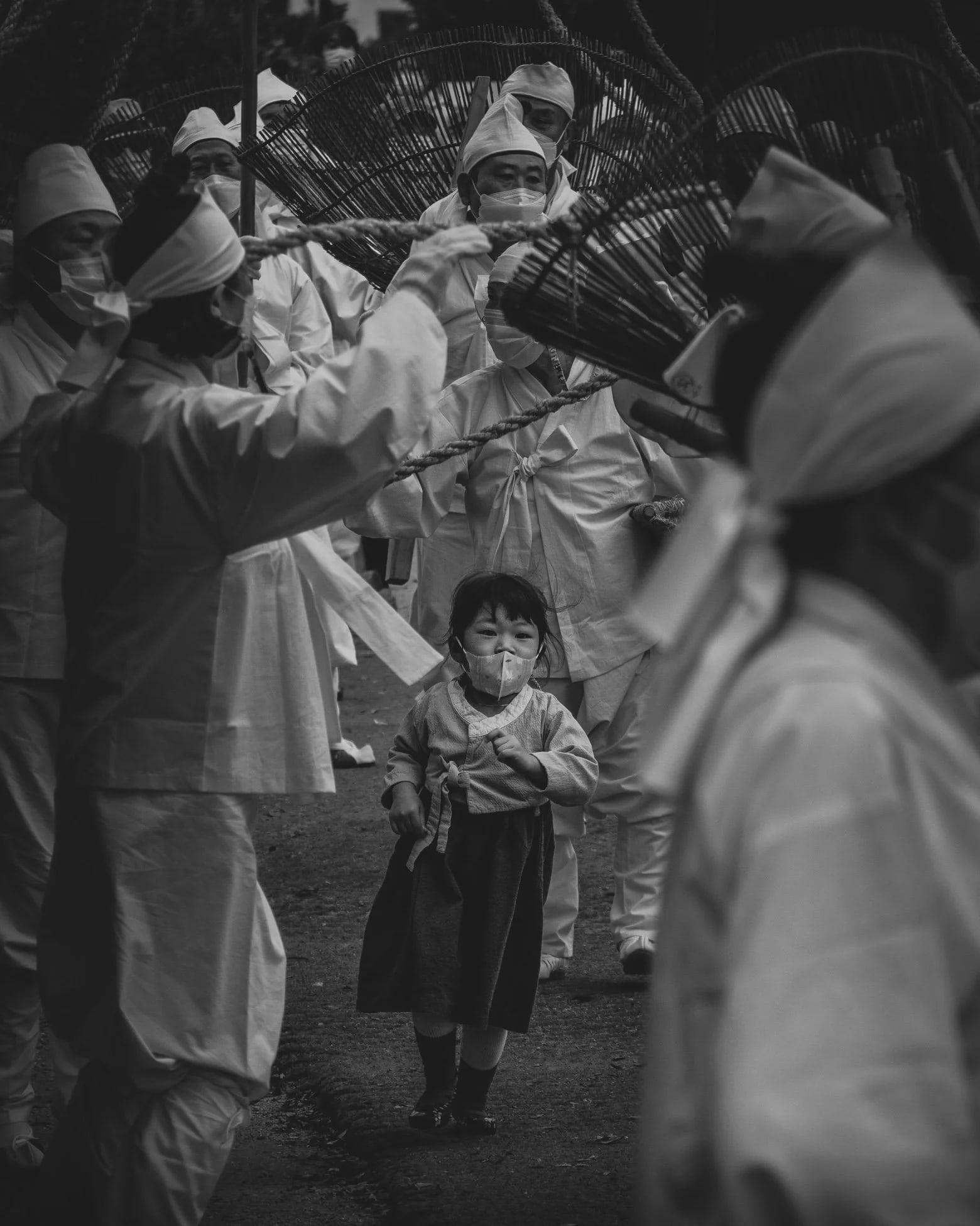 Child in traditional dress walking among adults in a procession, in black and white