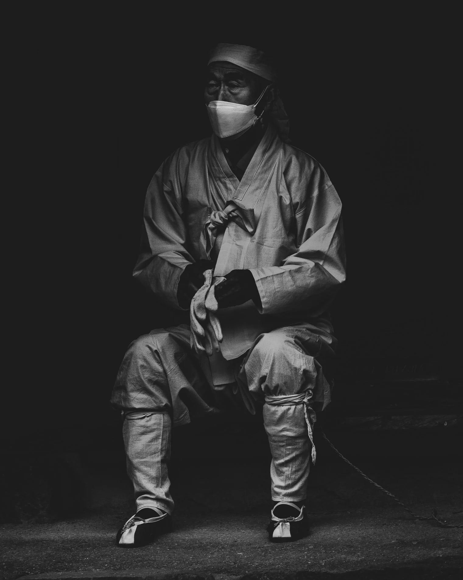 Man in traditional clothing and a face mask seated against a dark background
