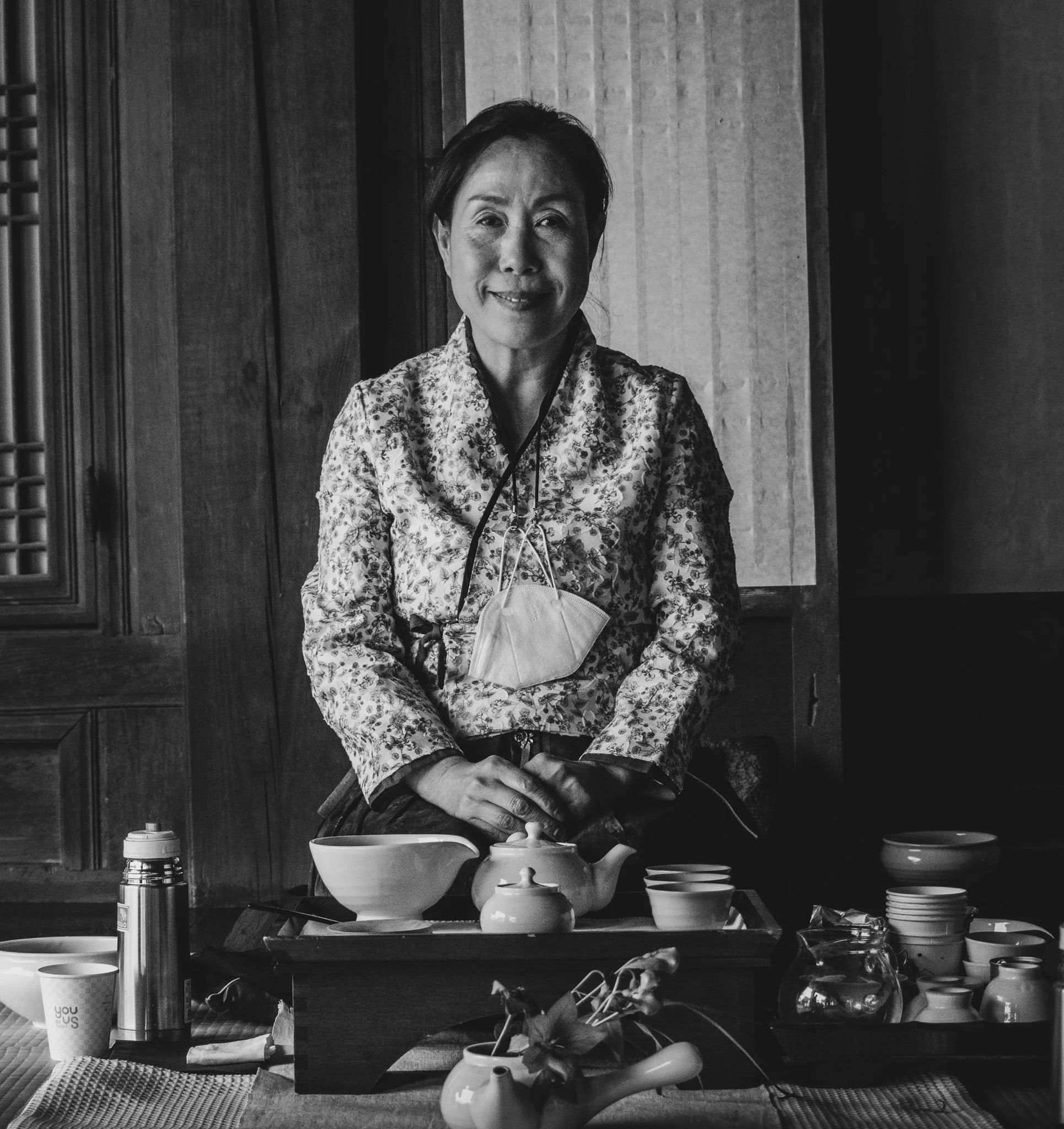 Black-and-white portrait of a woman in a patterned hanbok smiling behind a low tea table