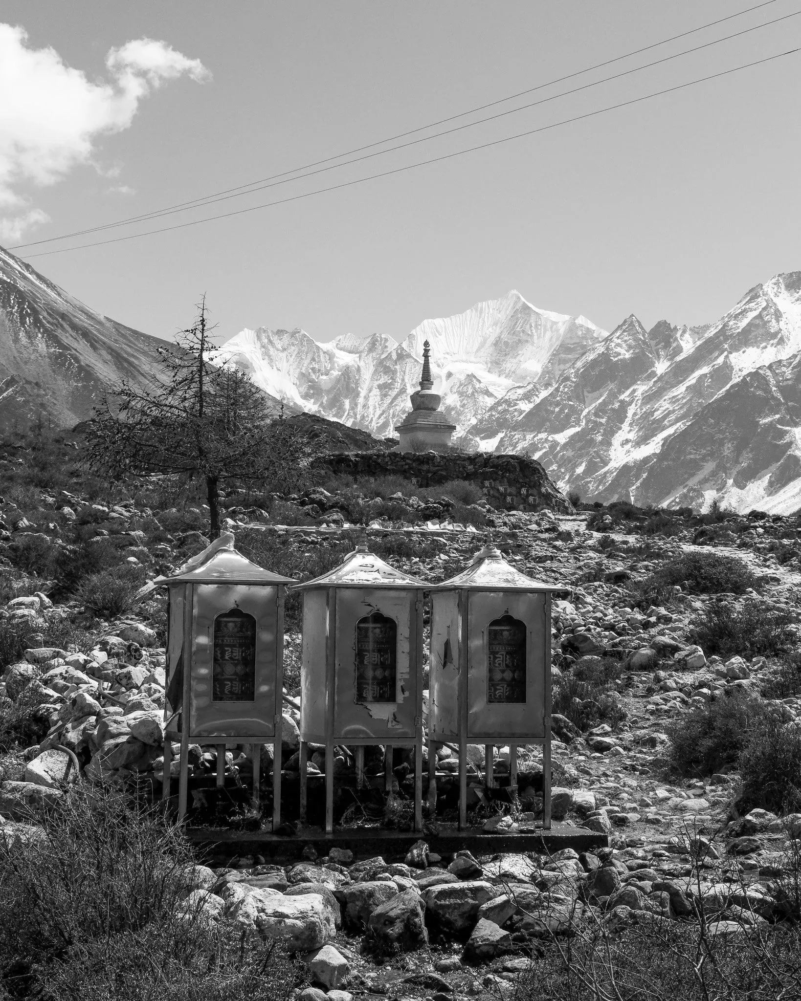 Black and white photo of small prayer wheel houses and a stupa with snow peaks behind, near Kyanjin Gompa