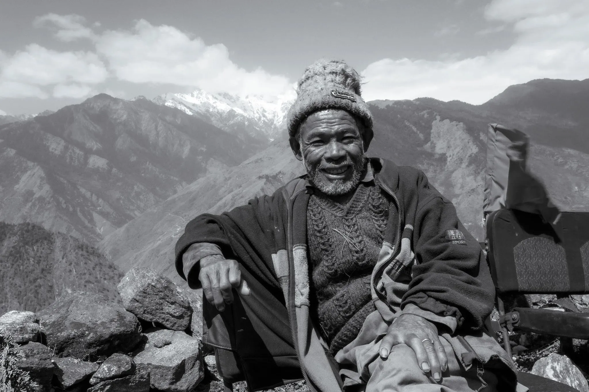 Black and white portrait of a smiling older man in a woollen hat with mountains behind