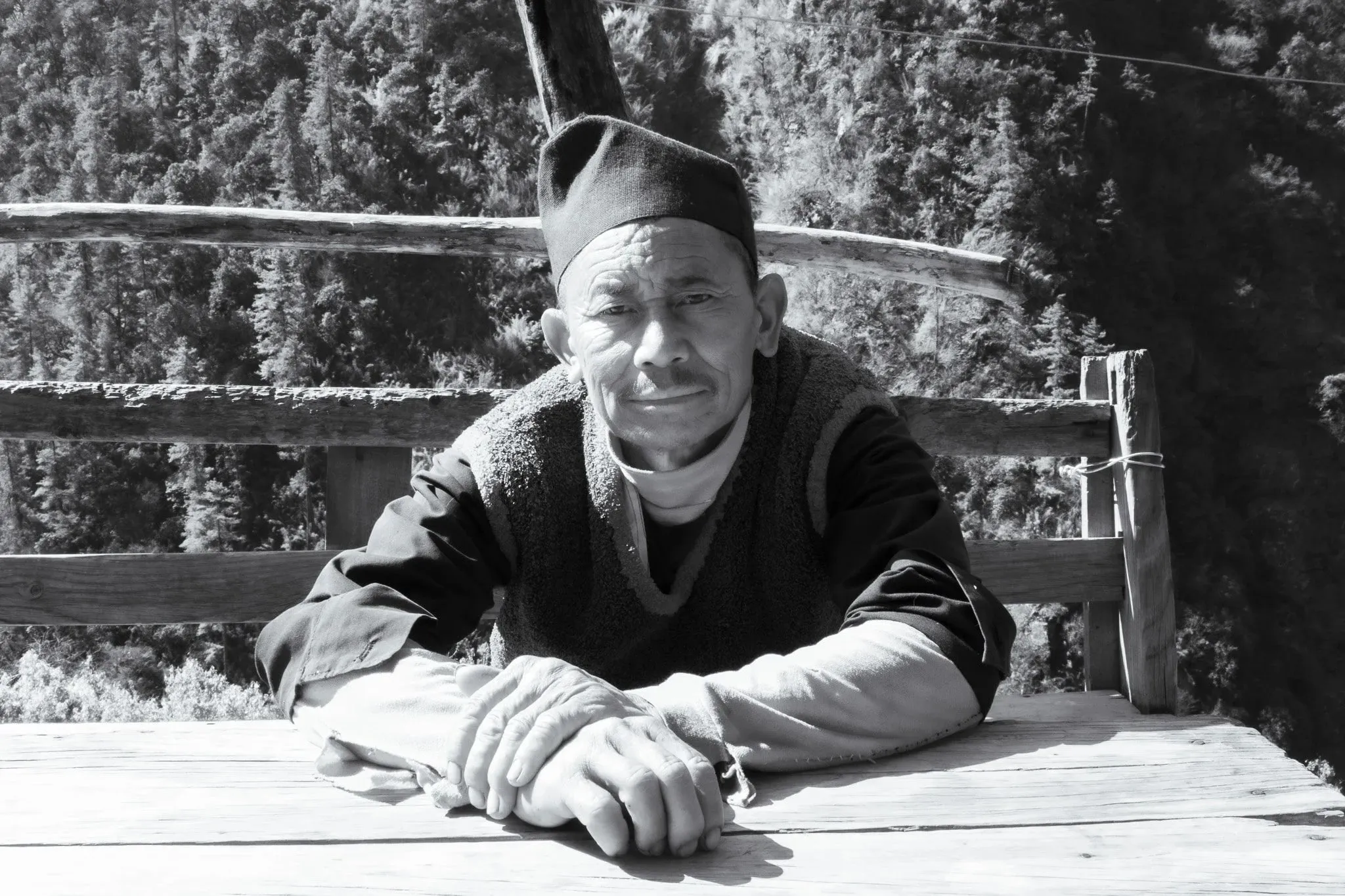 Black and white portrait of a man in a cap resting his arms on a wooden table