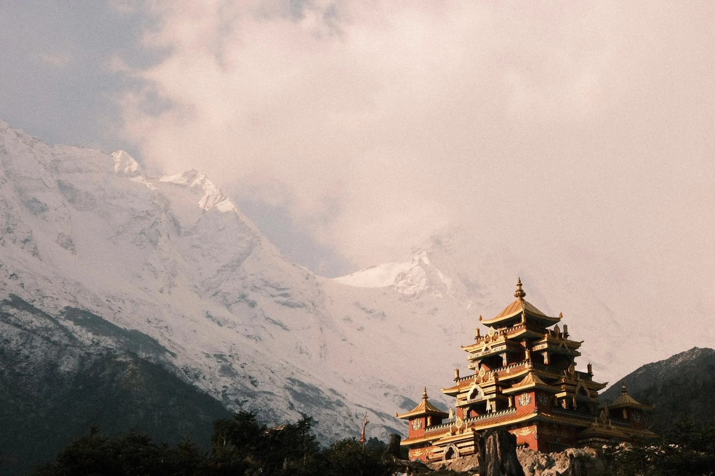 A monastery with tiered roofs beneath cloud-covered snow peaks near Lho