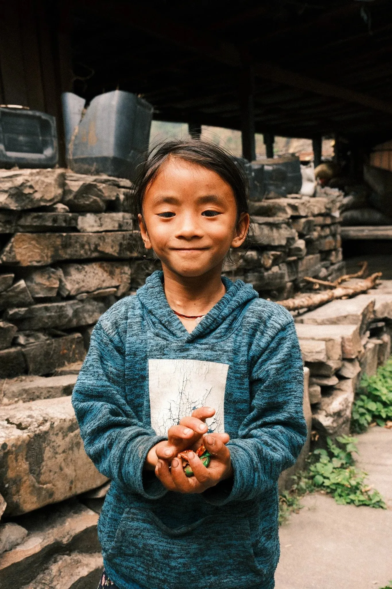 A smiling girl in a blue jumper holding something in her hands in front of a stone wall