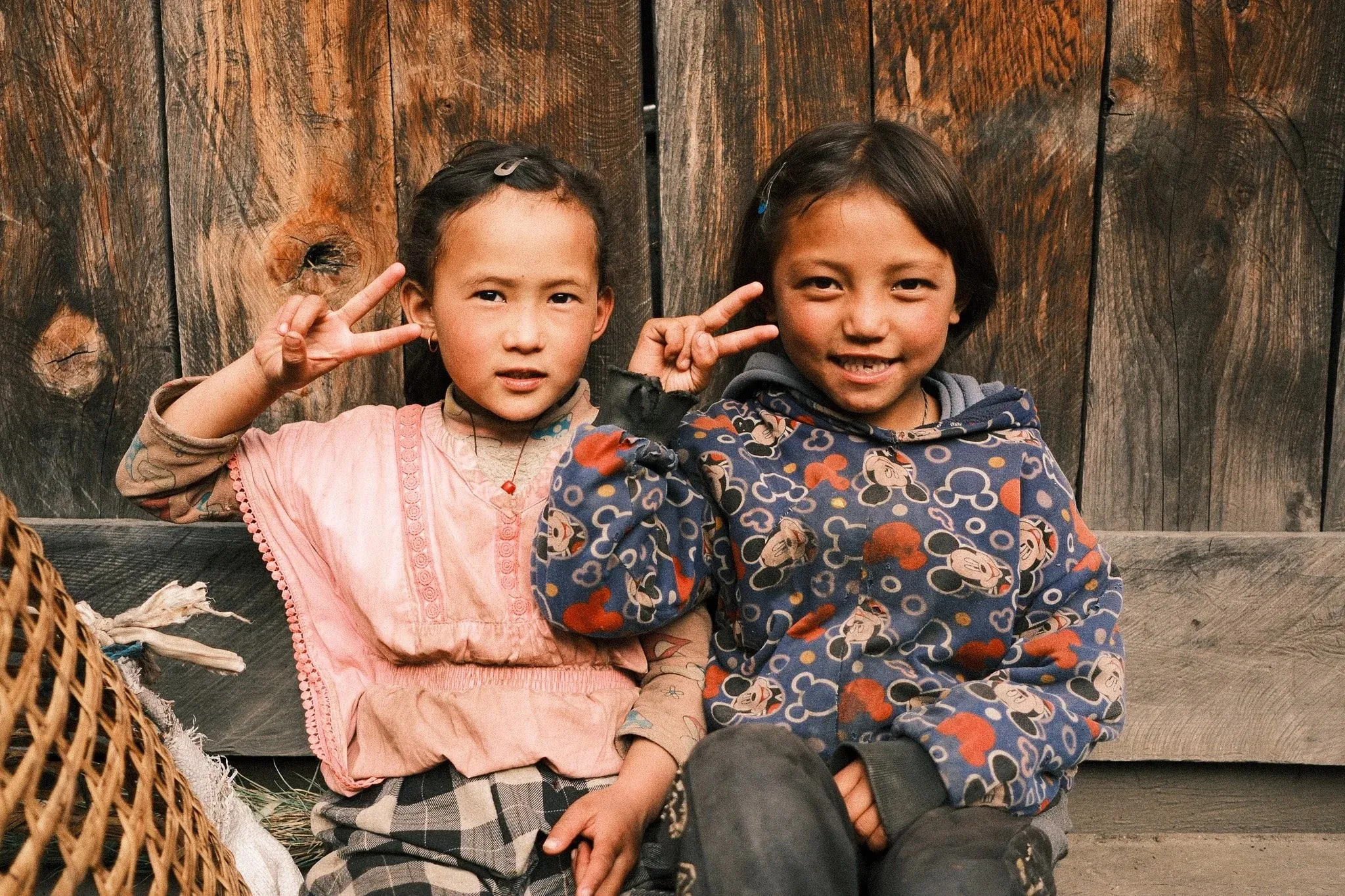 Two young girls making peace signs while sitting against a wooden wall