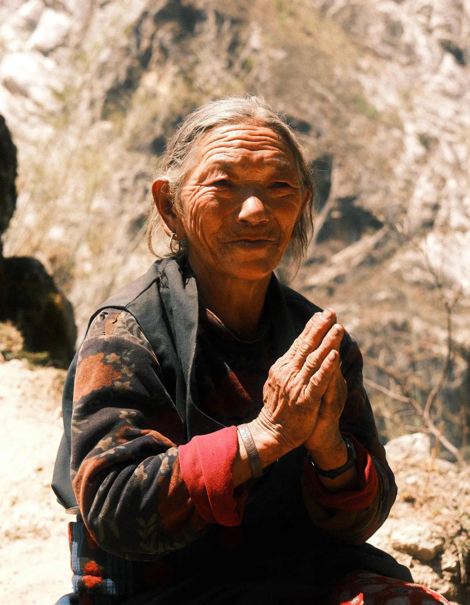 An older woman holding her hands together in greeting with a rocky slope behind