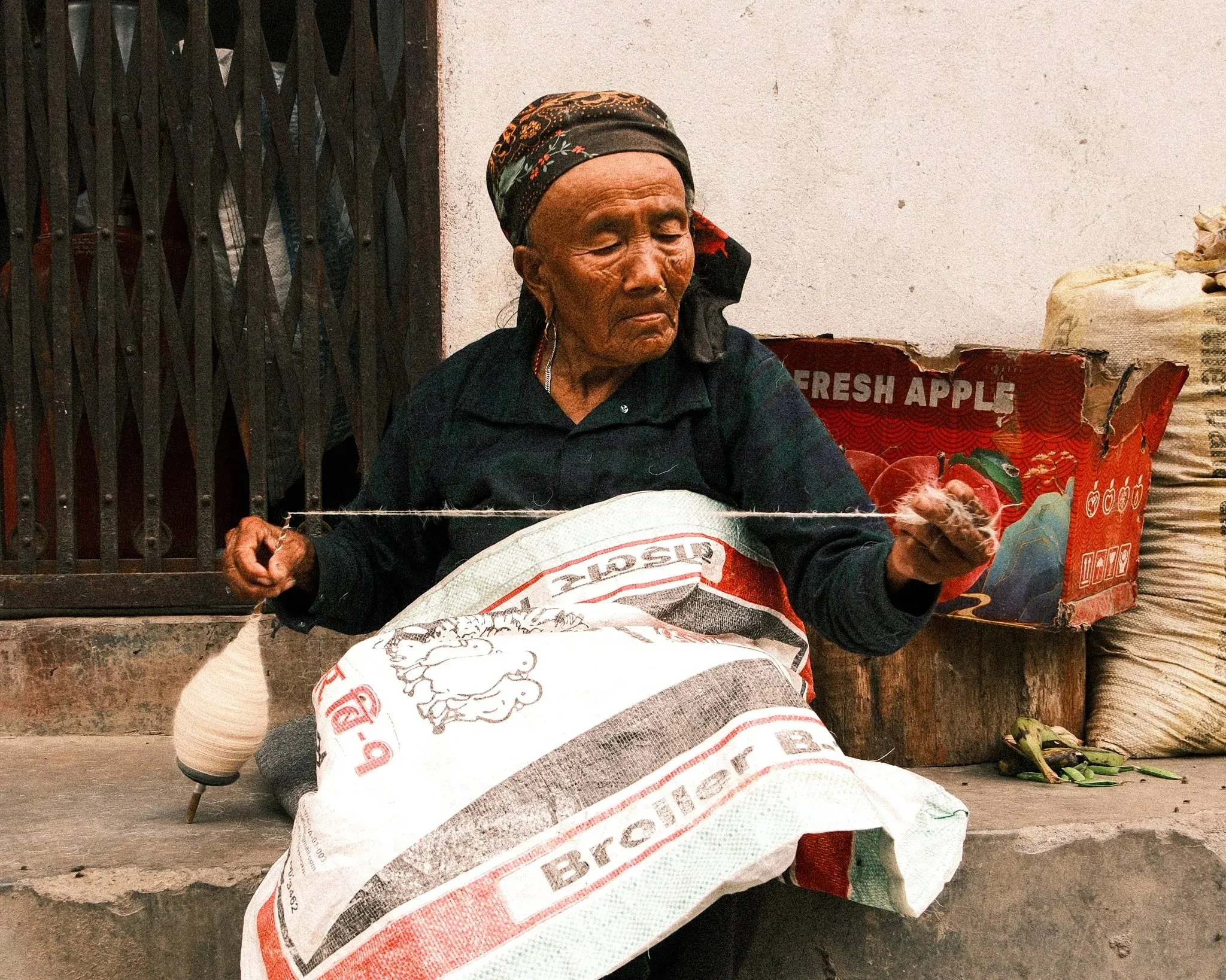 An older woman spinning wool with a drop spindle, sitting on a sack