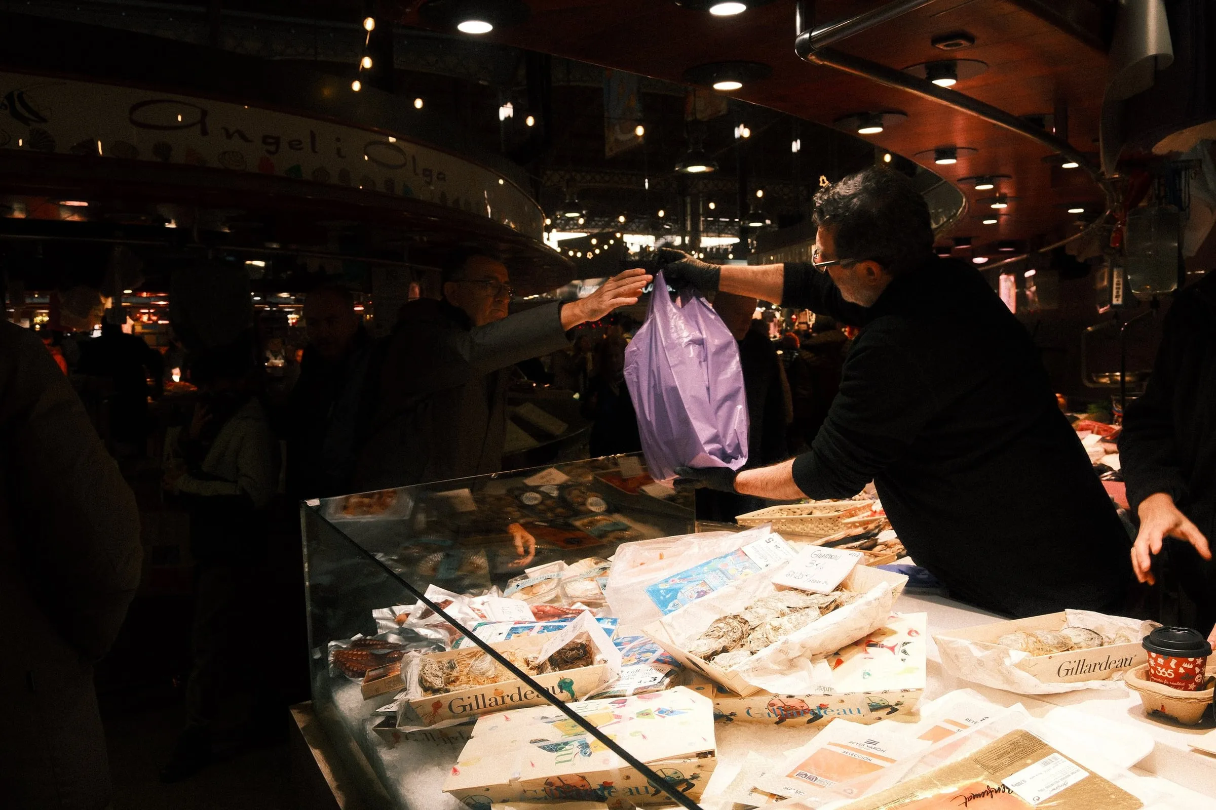 A stallholder hands a bag across a counter at a busy indoor market