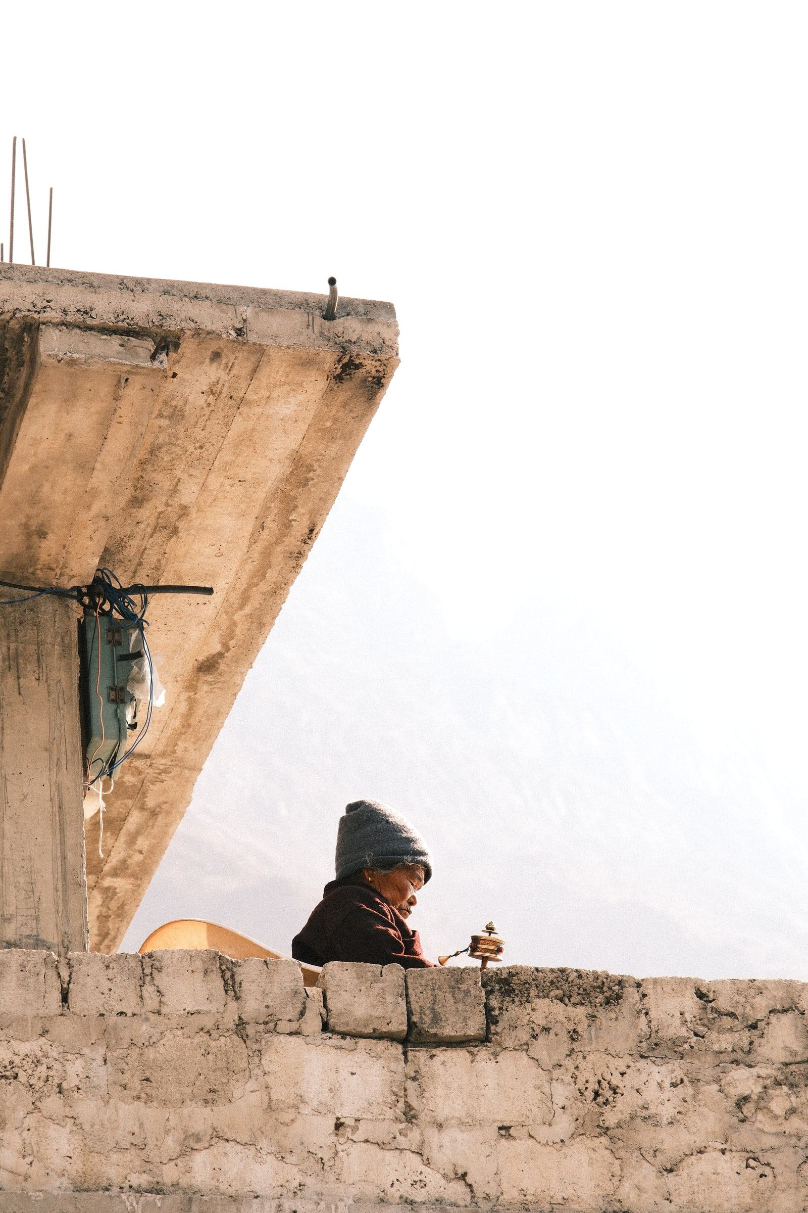 Person in a woollen hat looking out from behind a stone wall against a pale sky