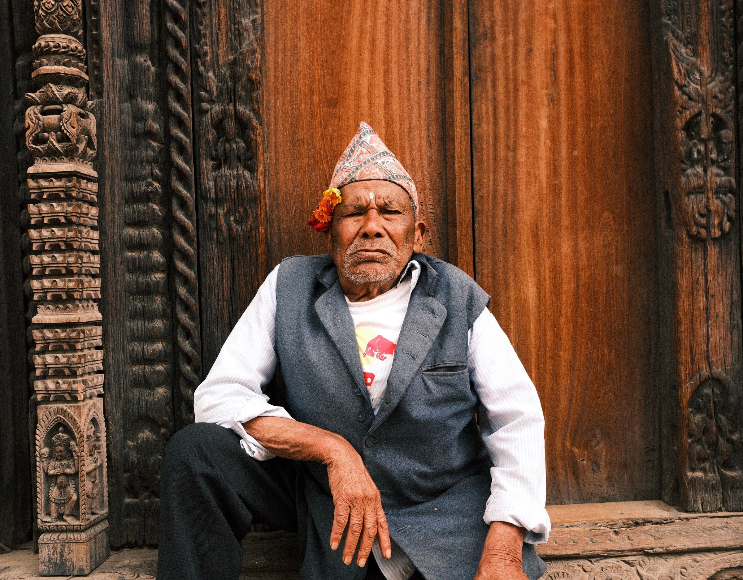 Older man in a dhaka topi and grey waistcoat sitting in front of carved wooden doors