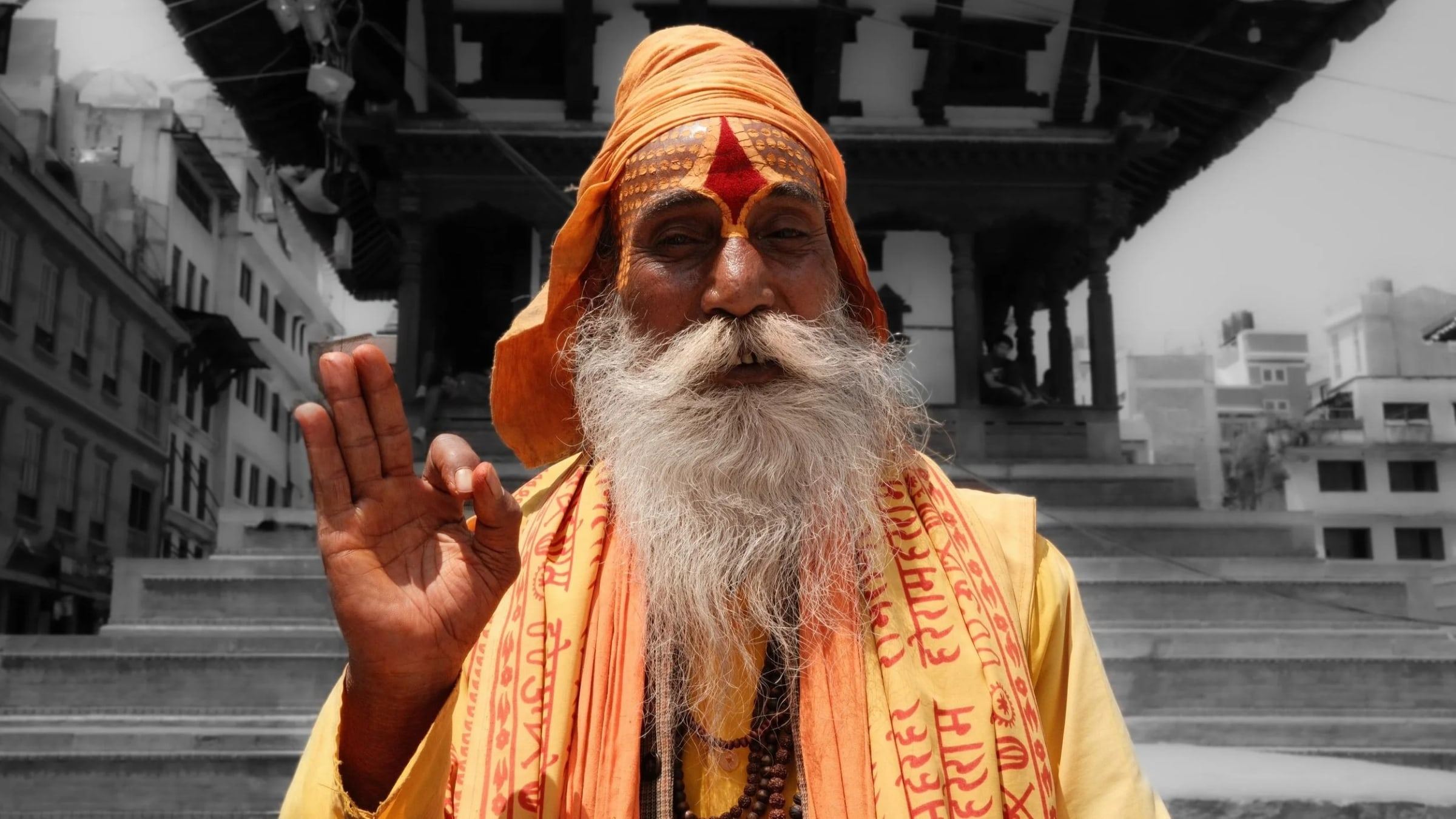 Sadhu with a long white beard and orange robes raising one hand in front of a temple