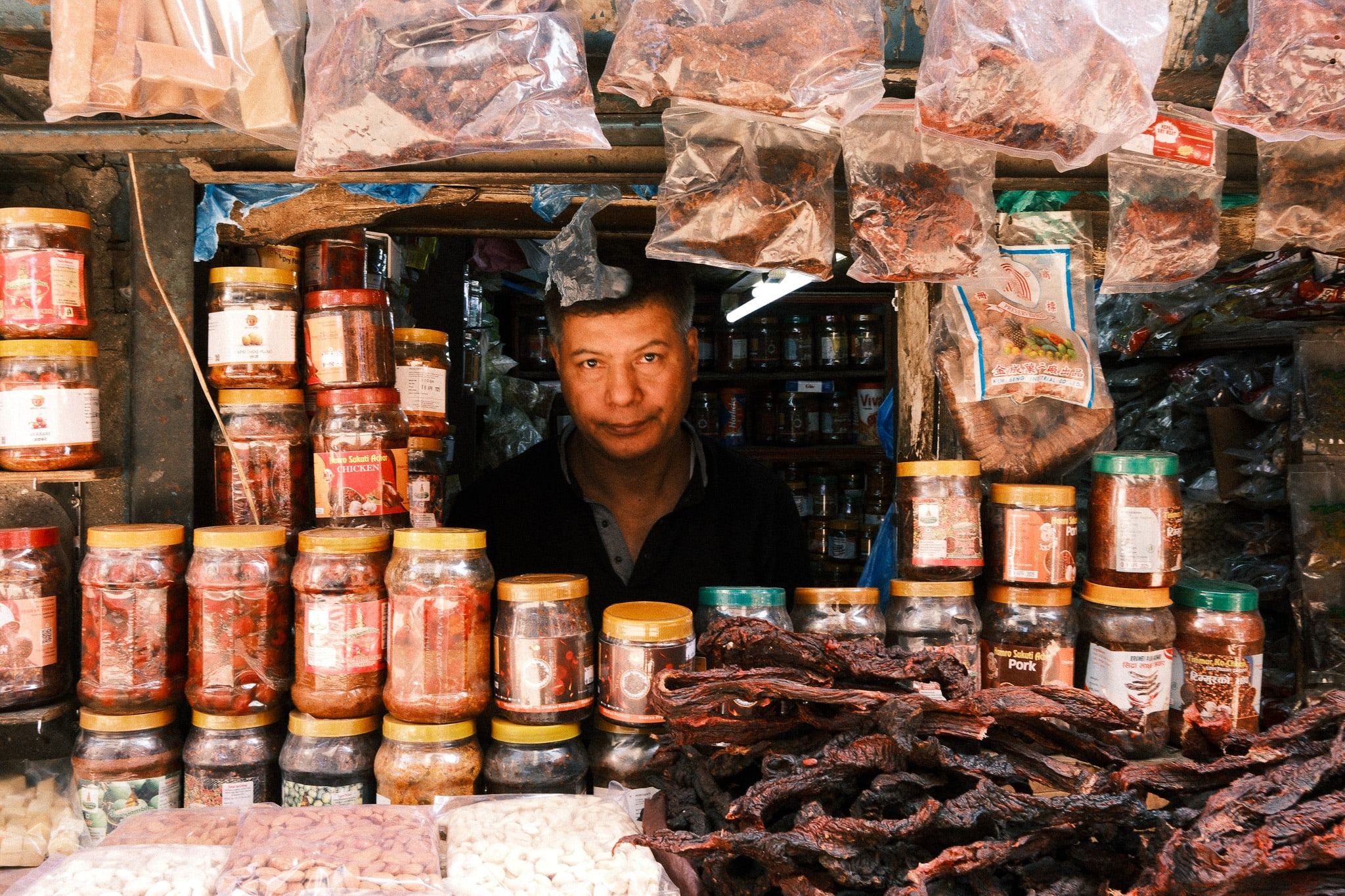 Shopkeeper framed by jars and hanging packets in a small shop