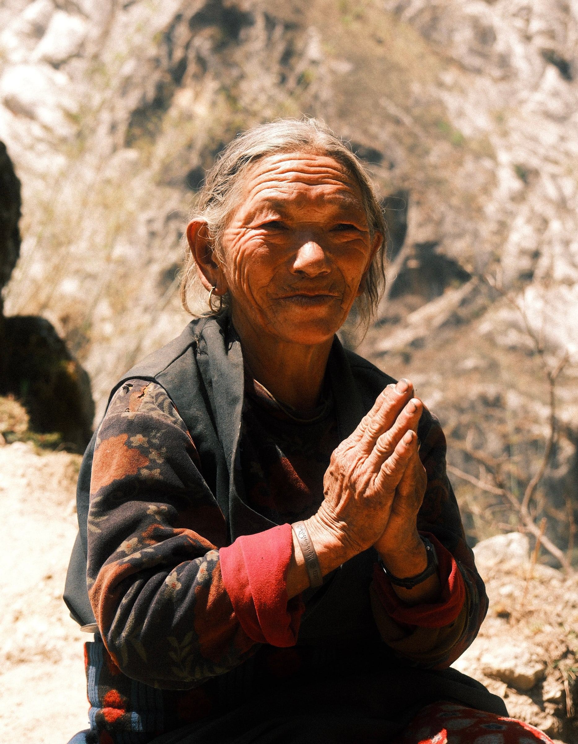 Older woman in a patterned jacket pressing her palms together on a rocky hillside