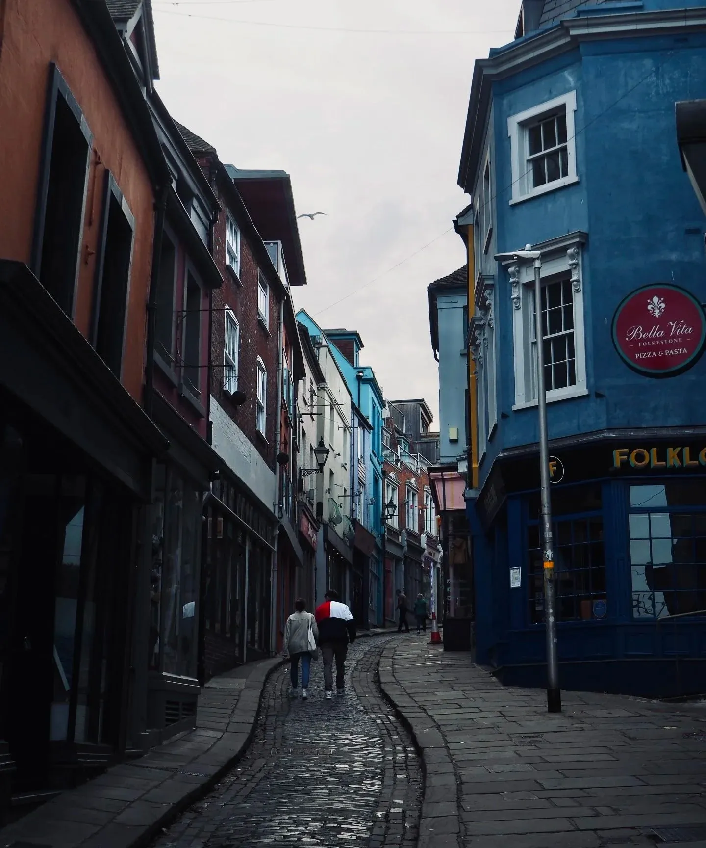 Two people walking up the wet cobbled Old High Street between colourful shopfronts